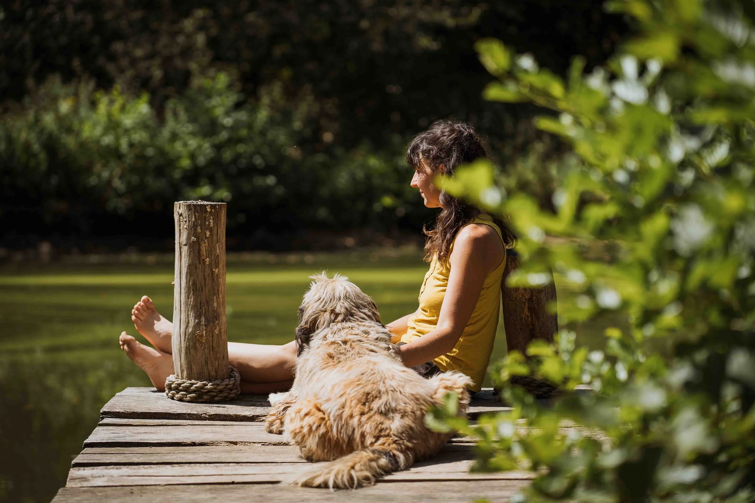 Jeune femme assise avec un chien, au bord de l'eau, sous des arbres, en plein air par une journée ensoleillée.