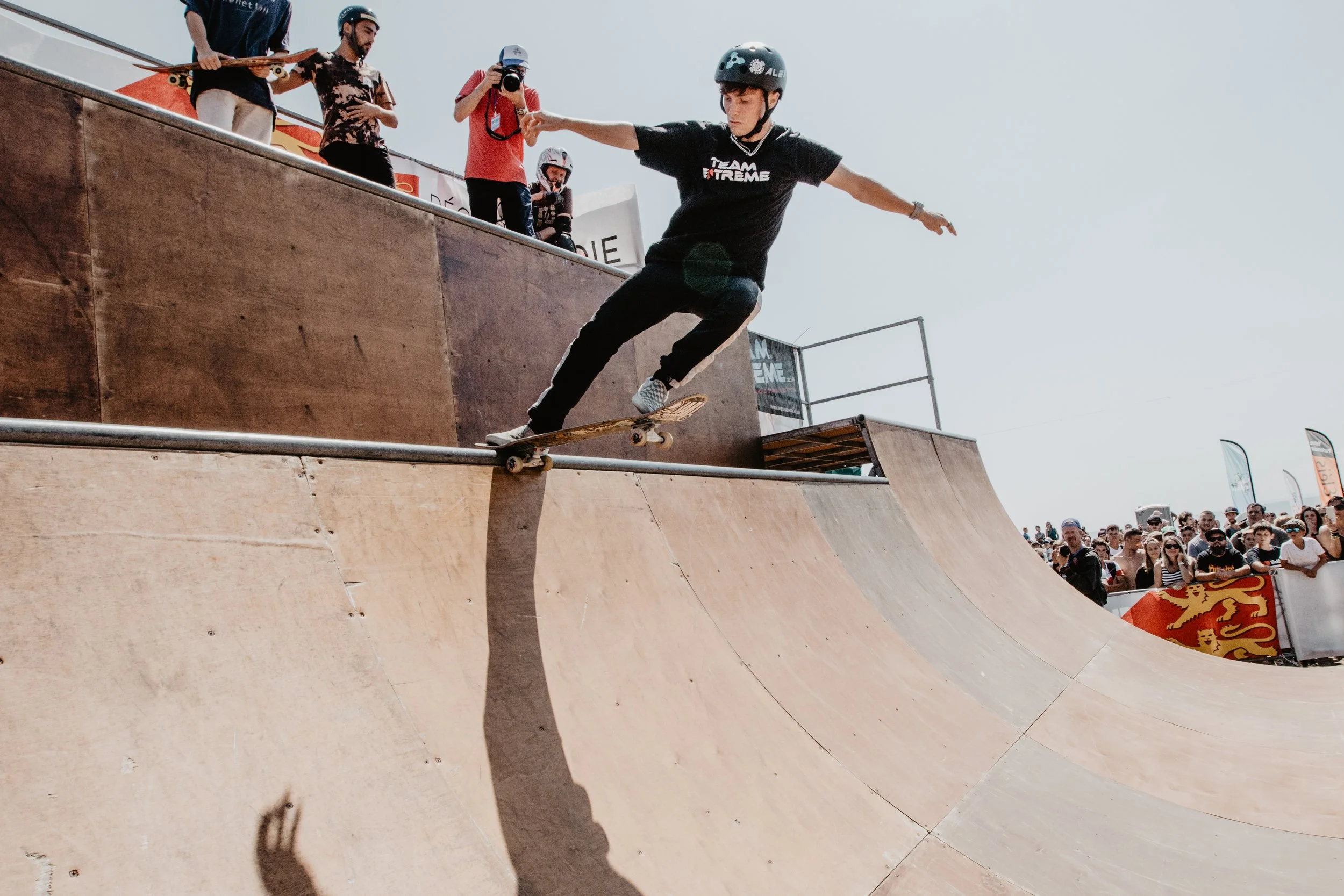 Un skateur en casque et T-shirt noir effectue une figure au skate sur une rampe en bois lors d'un événement de roller ou skateboard, avec une foule de spectateurs et plusieurs personnes en arrière-plan.