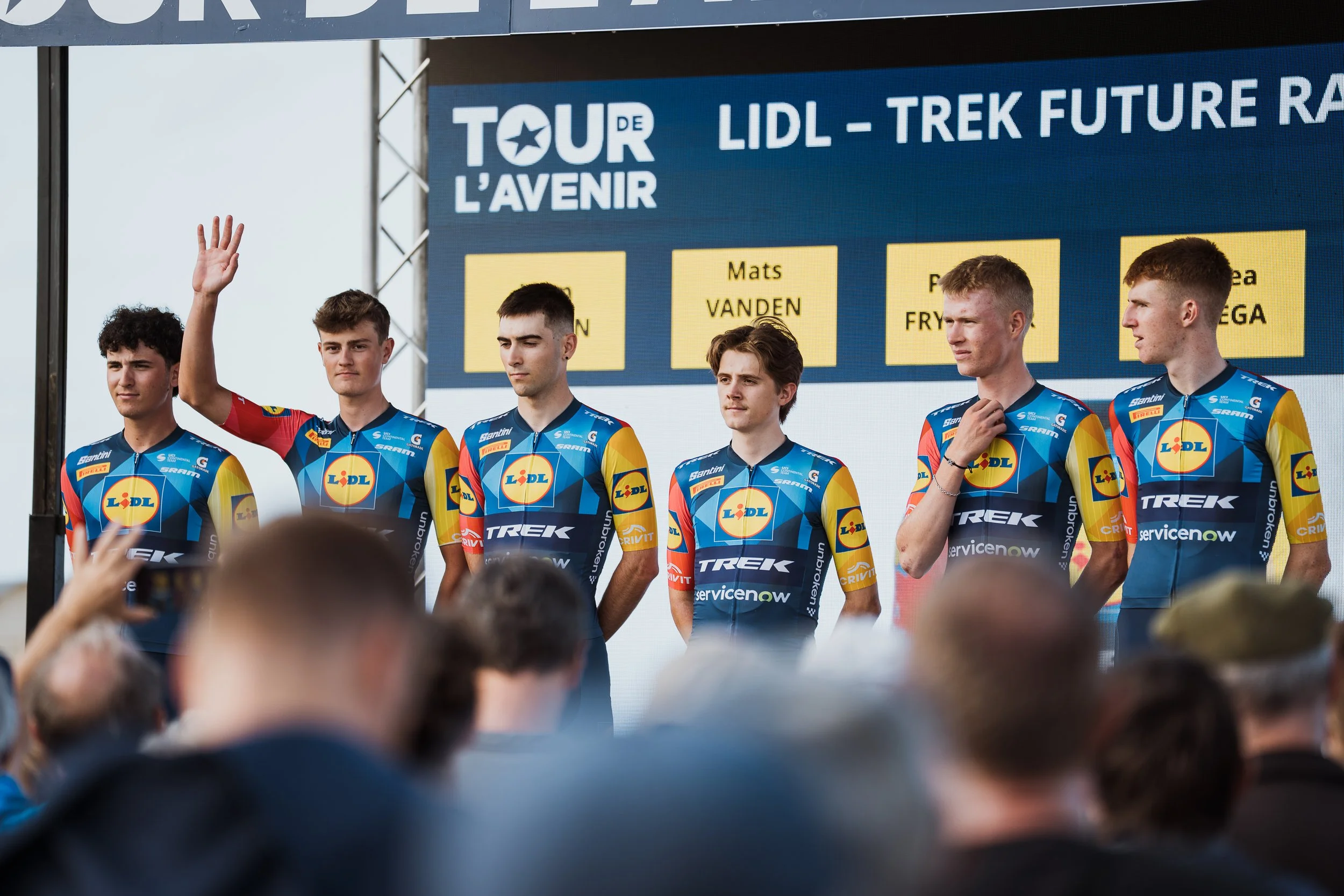 Un groupe de jeunes cyclistes en uniforme, en train de saluer un public lors d'une cérémonie ou d'une présentation de l'équipe. En arrière-plan, un écran affiche des informations sur la compétition ou l'événement, avec le texte 'Tour de l'avenir', 'Lidl - Trek Future', et des noms de sponsors ou de participants.