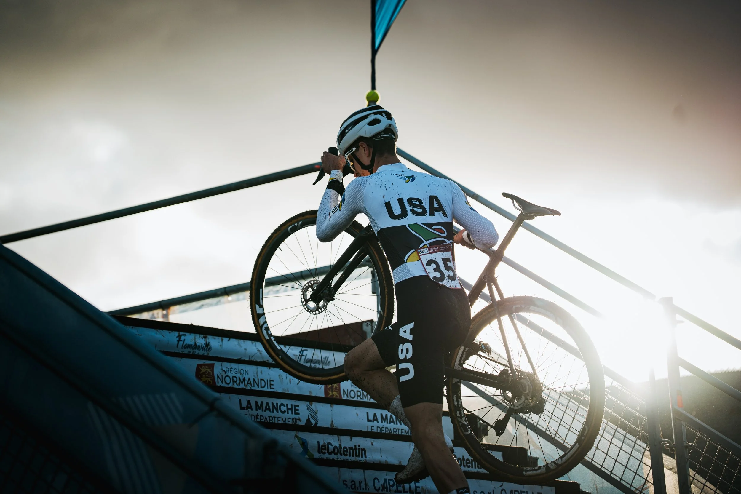 Coureur cycliste portant un maillot avec l'inscription 'USA' et un casque, portant un vélo en le tenant, lors d'une compétition. La scène est prise en contre-plongée avec un ciel nuageux en arrière-plan.