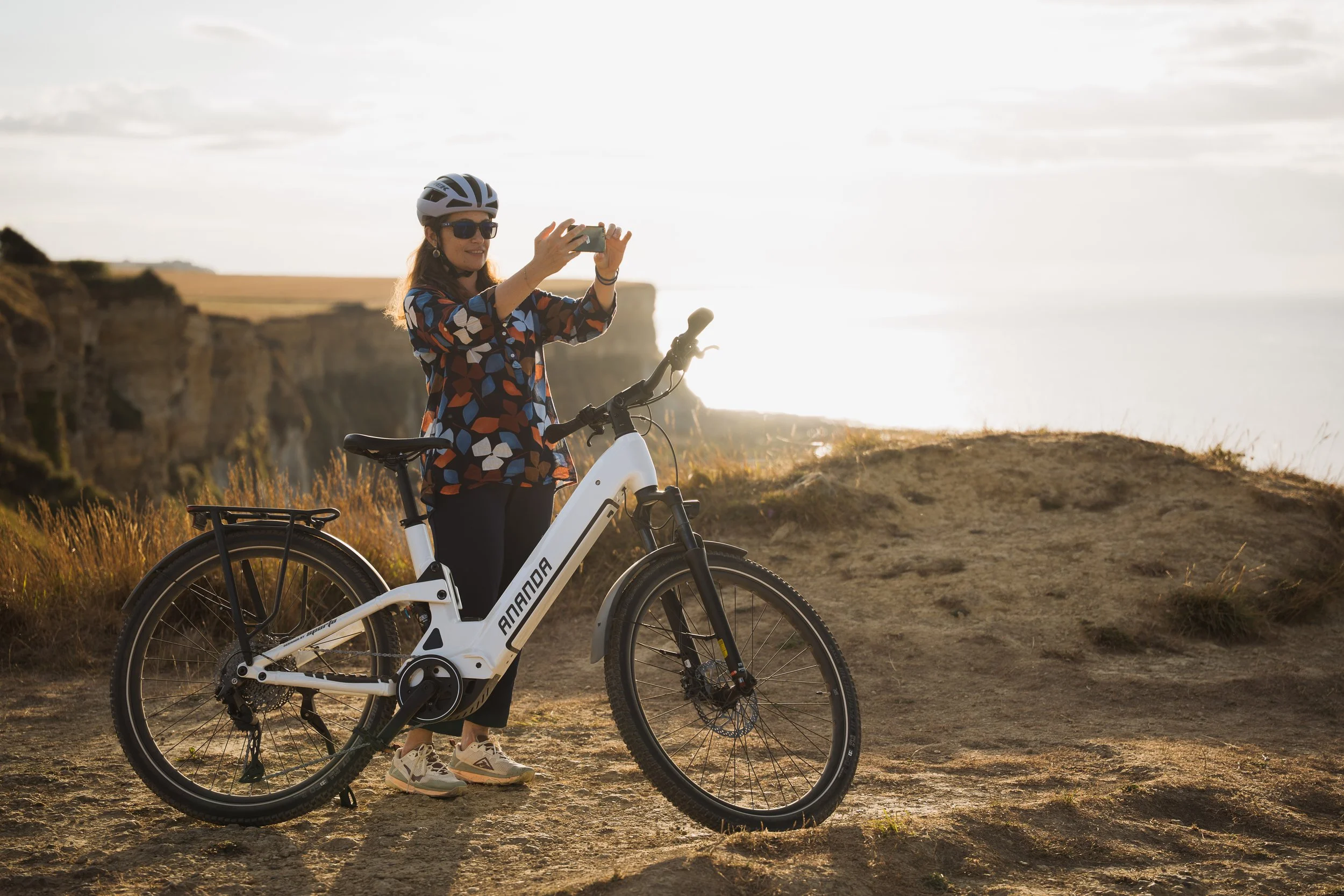 Femme en chemise à motifs, portant un casque et des lunettes de soleil, prenant une photo avec son téléphone tout en se tenant à côté de son vélo électrique sur un chemin côtier au coucher du soleil.