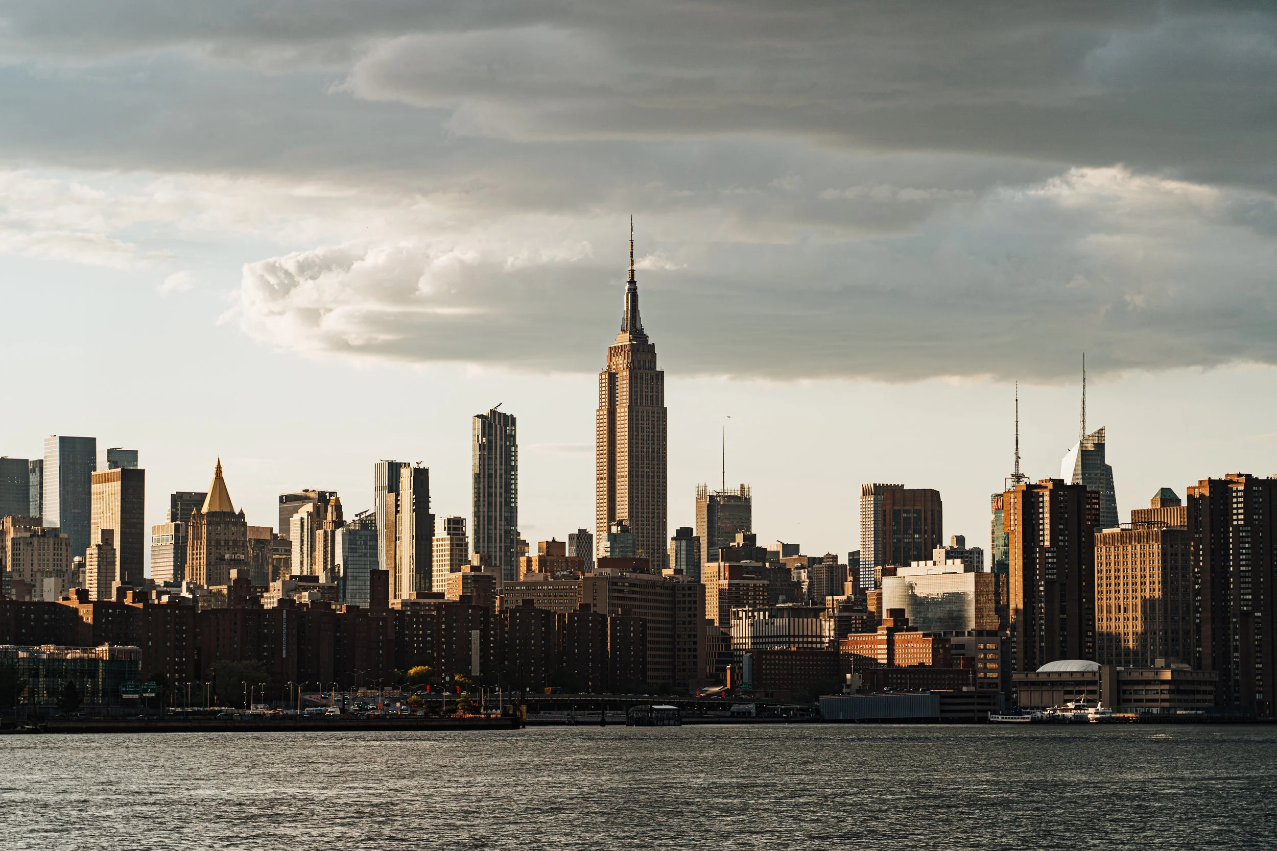 La silhouette de la skyline de New York avec des gratte-ciel emblématiques, notamment l'Empire State Building, sous un ciel nuageux, vue depuis le fleuve.