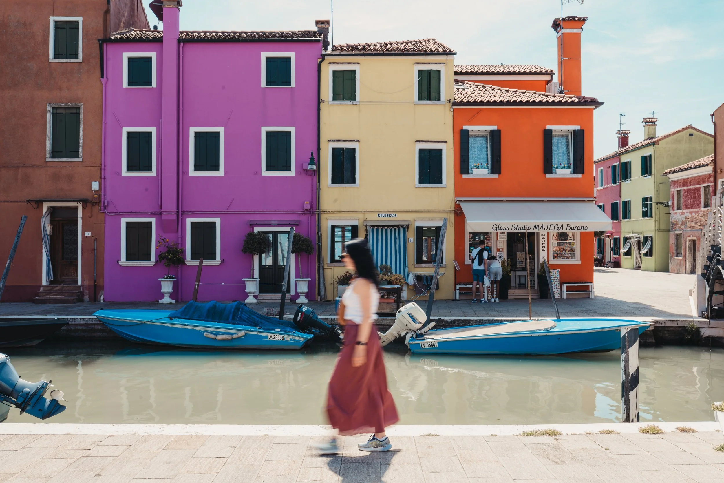 Des maisons colorées en vert, violet, jaune et orange avec des fenêtres et des terrasses, situées le long d'un canal avec des bateaux, un homme avec un sac à dos. Des personnes marchent sur le trottoir.