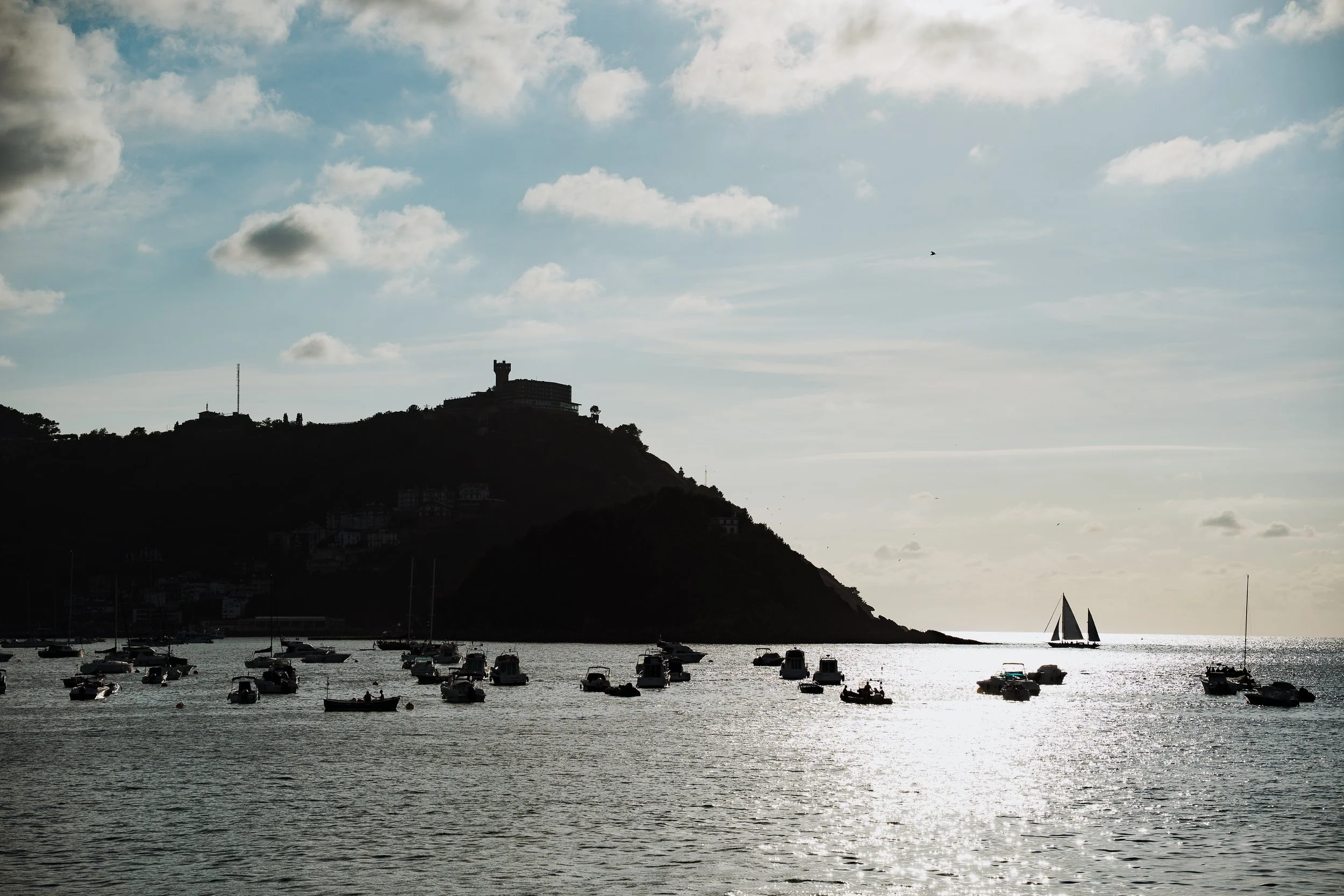 Vue sur une baie avec des bateaux au premier plan, une colline avec un bâtiment en silhouette et un château au sommet, ciel partiellement nuageux, un voilier blanc naviguant dans l'eau.
