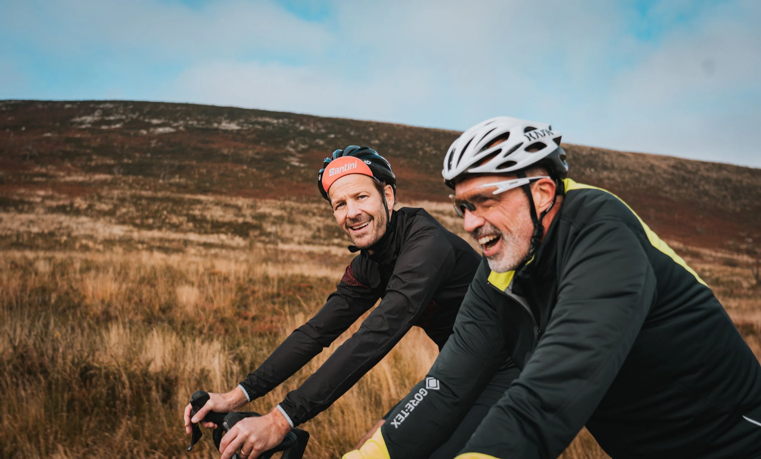 Deux hommes en tenues de cyclisme portant des casques en pleine nature, souriant lors d'une sortie à vélo dans un paysage de collines avec de l'herbe sèche.