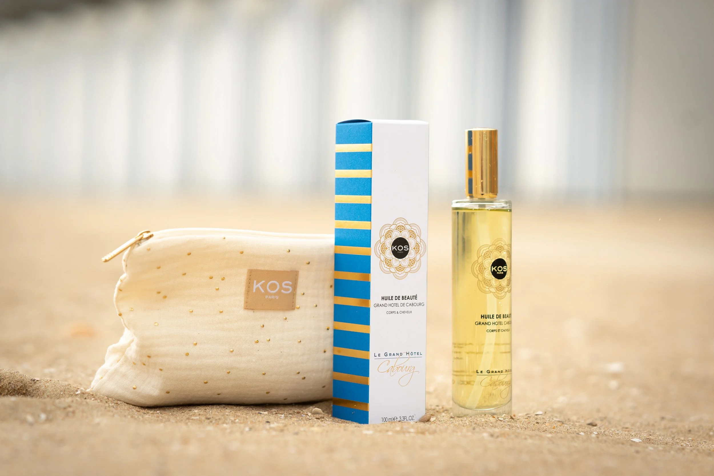 Une bouteille d'huile de beauté, un boîte et une pochette beige en tissu avec le logo KOS Paris, sur une plage.