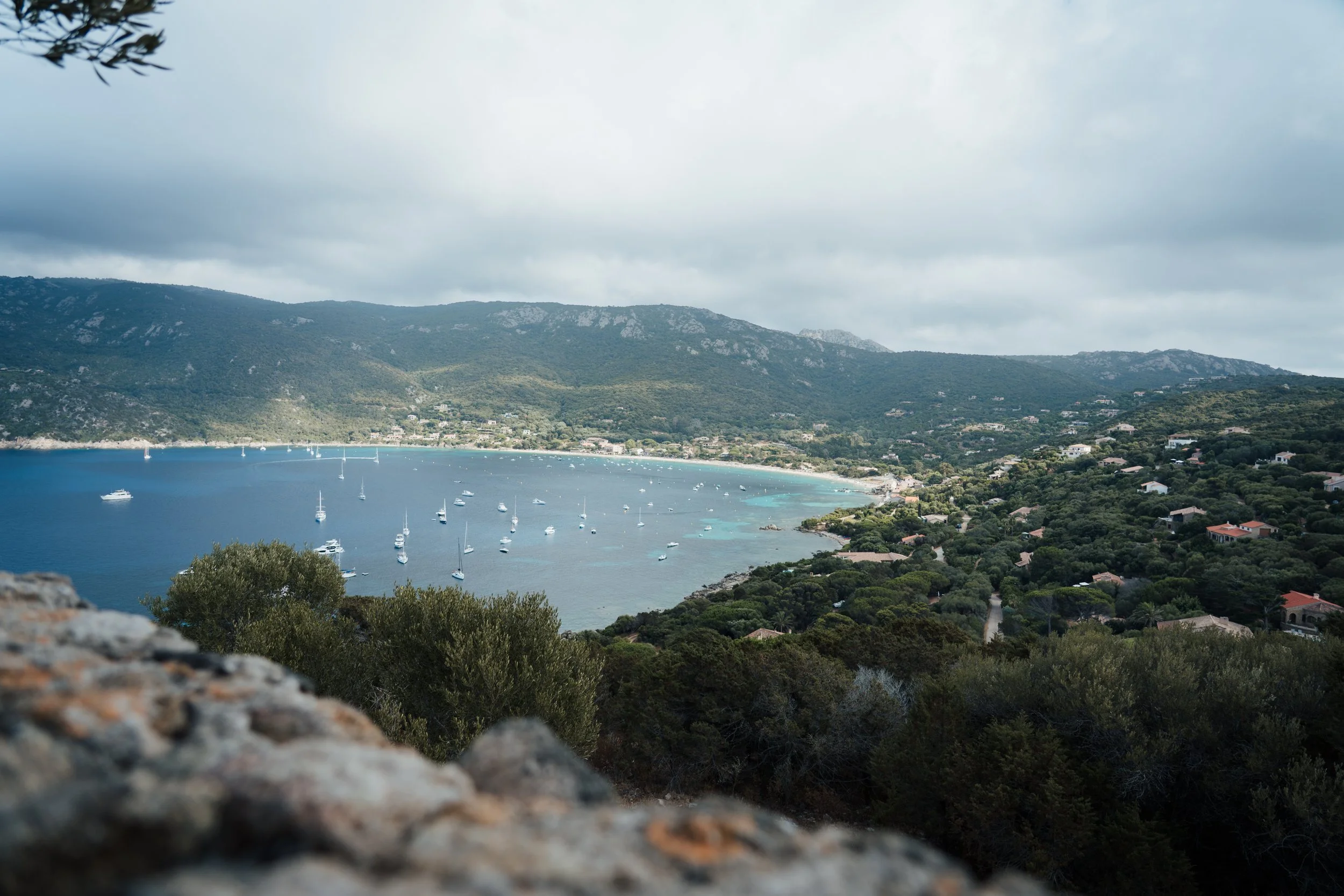 Vue d'une baie avec des bateaux à voile dans l'eau calme, entourée de montagnes vertes et de petites maisons, sous un ciel nuageux.