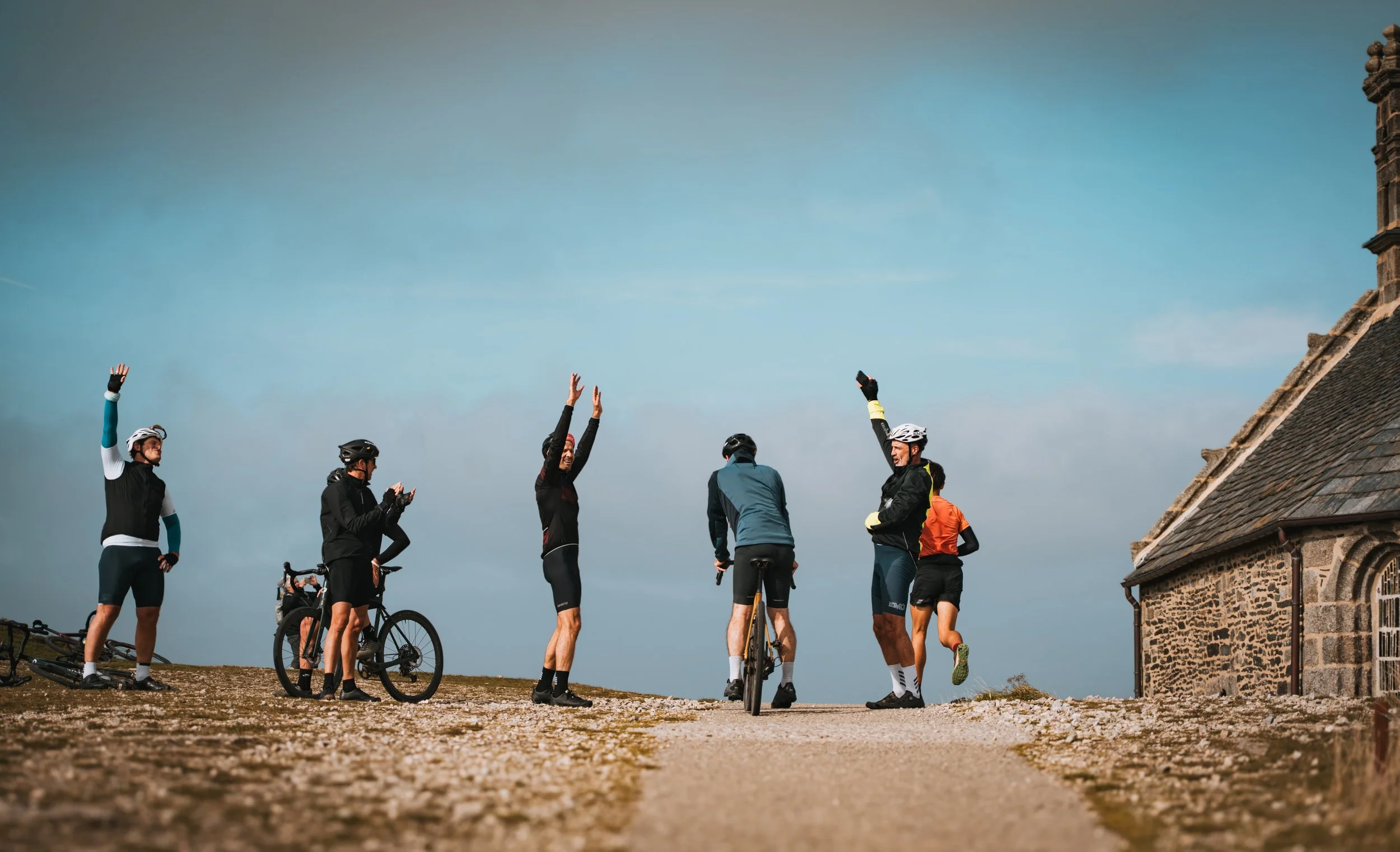 Groupe de cyclistes en tenue de sport se saluant ou discutant à l'extérieur, près d’un bâtiment ancien en pierre, par une journée ensoleillée.