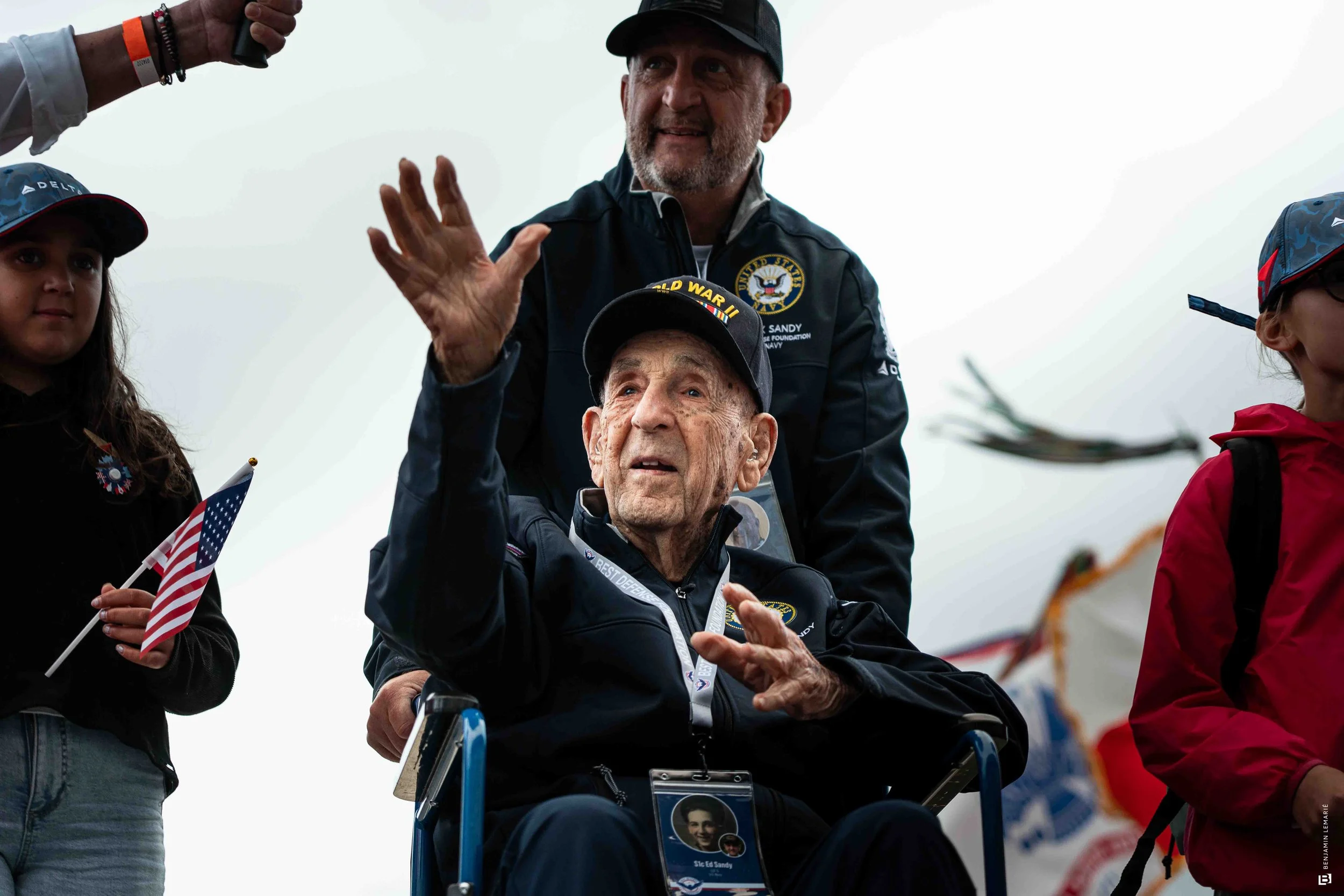 Un homme en fauteuil roulant entouré de jeunes et d'adultes, certains portant des casquettes et tenues militaires, lors d'un événement extérieur avec des drapeaux américains en main.