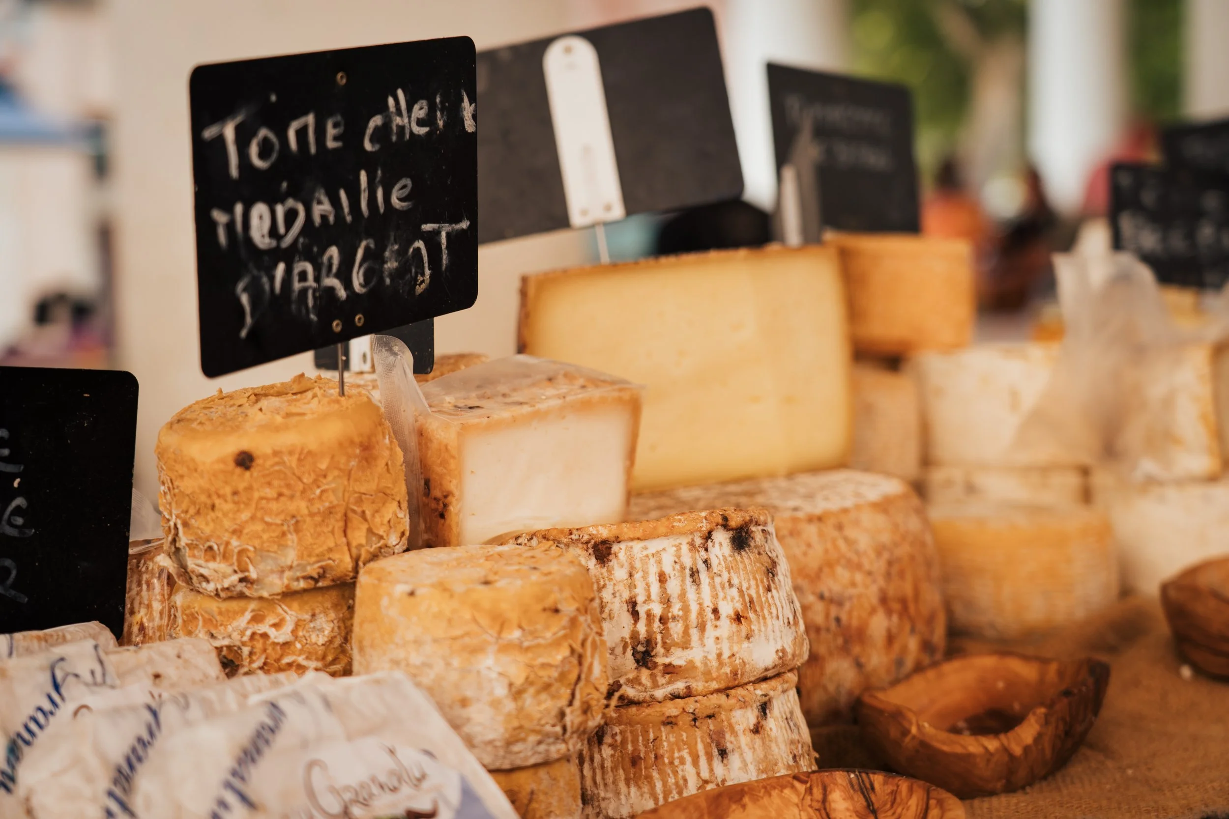 Différents fromages présentés en rangée avec des étiquettes en ardoise au-dessus, à l'intérieur d'une fromagerie ou d'un marché.