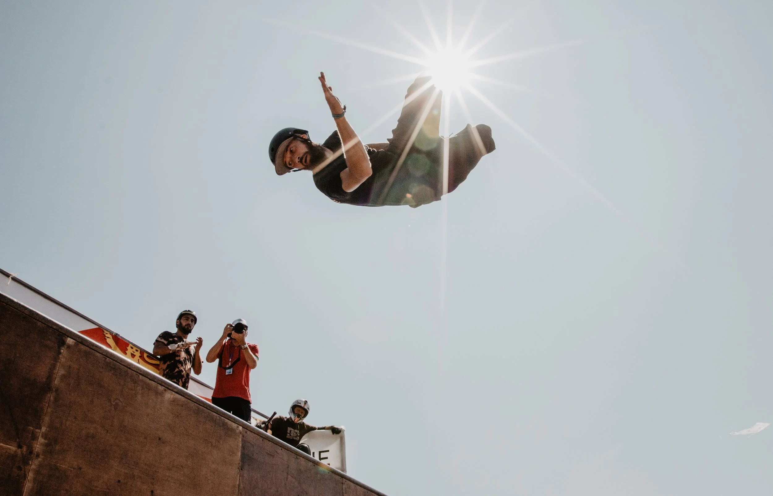 Une personne effectue une figure de skateboard ou de BMX dans un skatepark sous un soleil brillant dans un ciel dégagé. Deux autres personnes regardent et prennent des photos ou vidéo depuis le côté.
