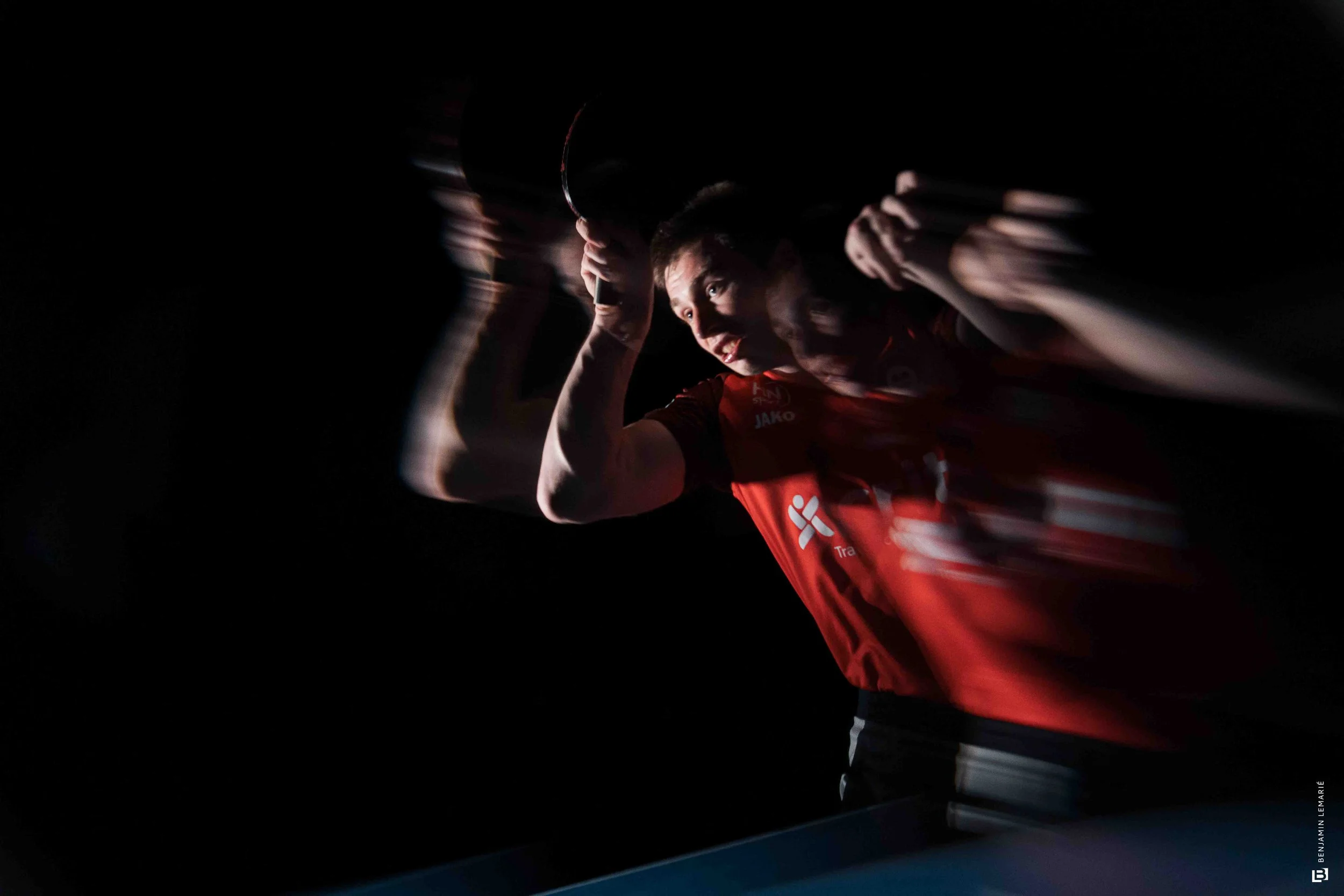 Jeune homme souriant en train de jouer au tennis, tenant une raquette dans une ambiance sombre avec un éclairage dramatique.