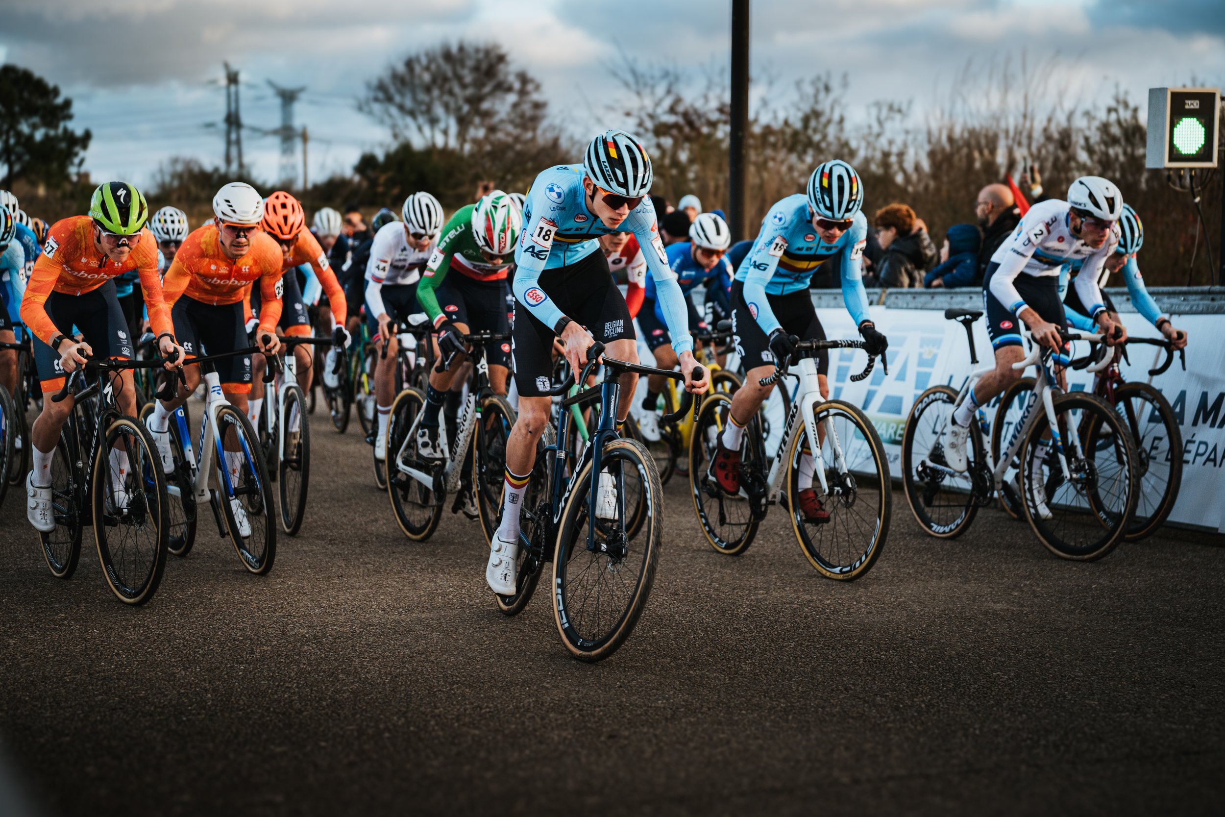 Cyclistes en compétition lors d'une course, portant des maillots colorés et casques, positionnés en aileron sur la ligne de départ, avec des spectateurs en arrière-plan et un feu vert indiquant le début de la course.