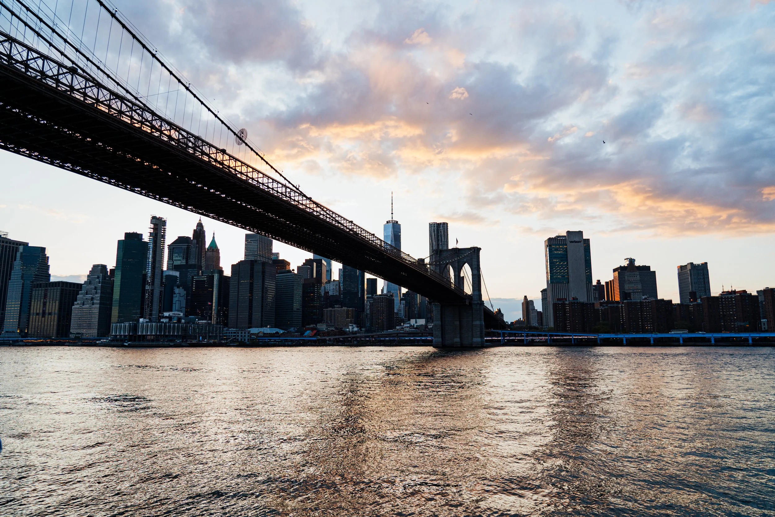Une vue de la skyline de New York avec le pont de Brooklyn au premier plan, le tout au coucher du soleil avec un ciel partiellement nuageux.