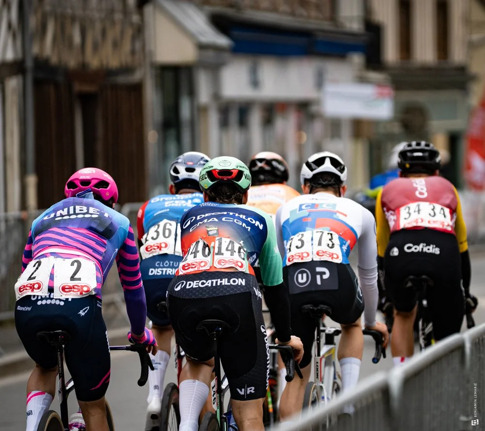 Groupe de cyclistes en compétition vue de derrière, portant des casques et des tenues de course colorées, roulant dans une rue urbaine.