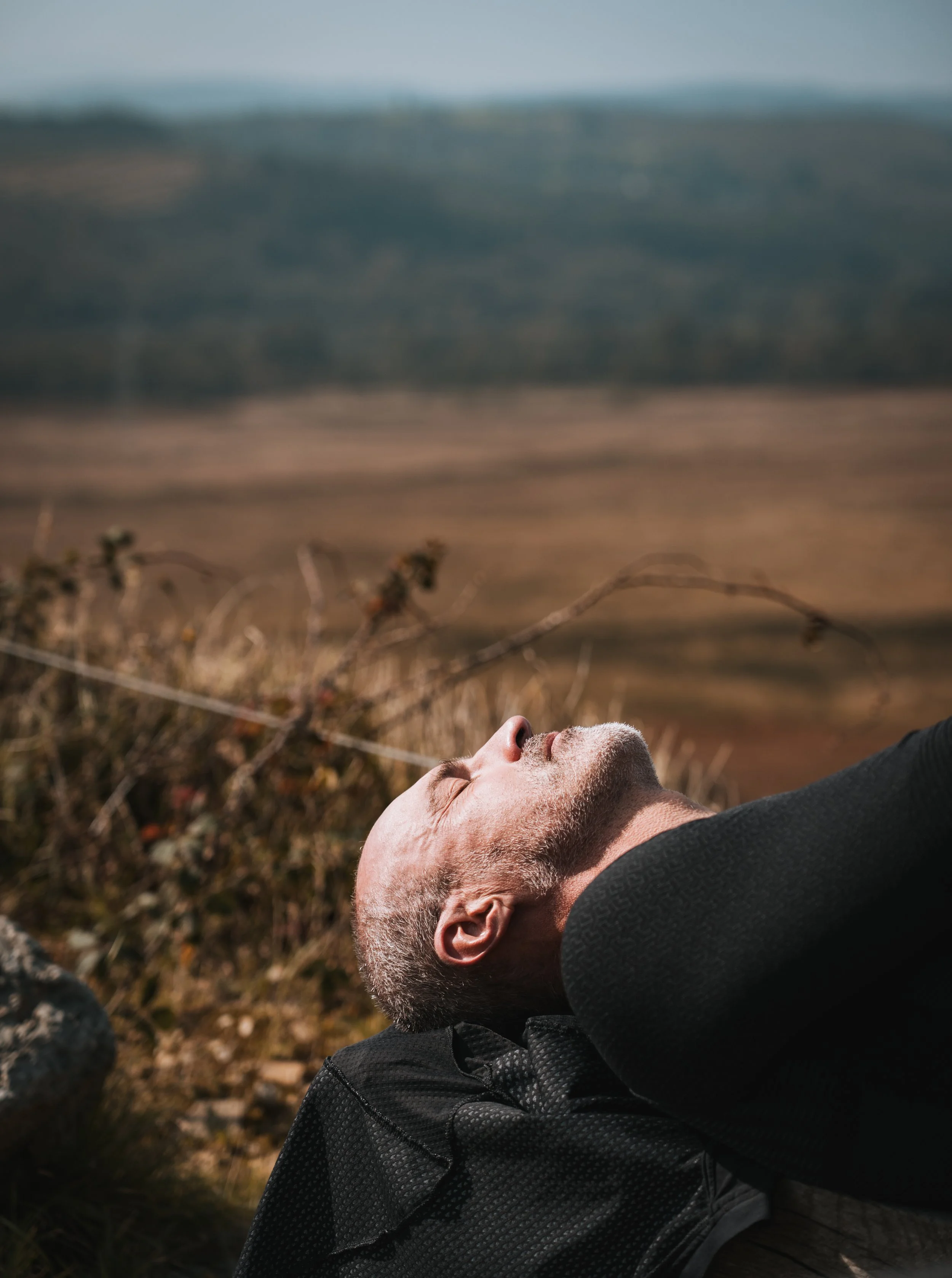Un homme allongé sur l'herbe, les yeux fermés, dans un paysage naturel avec des montagnes en arrière-plan.