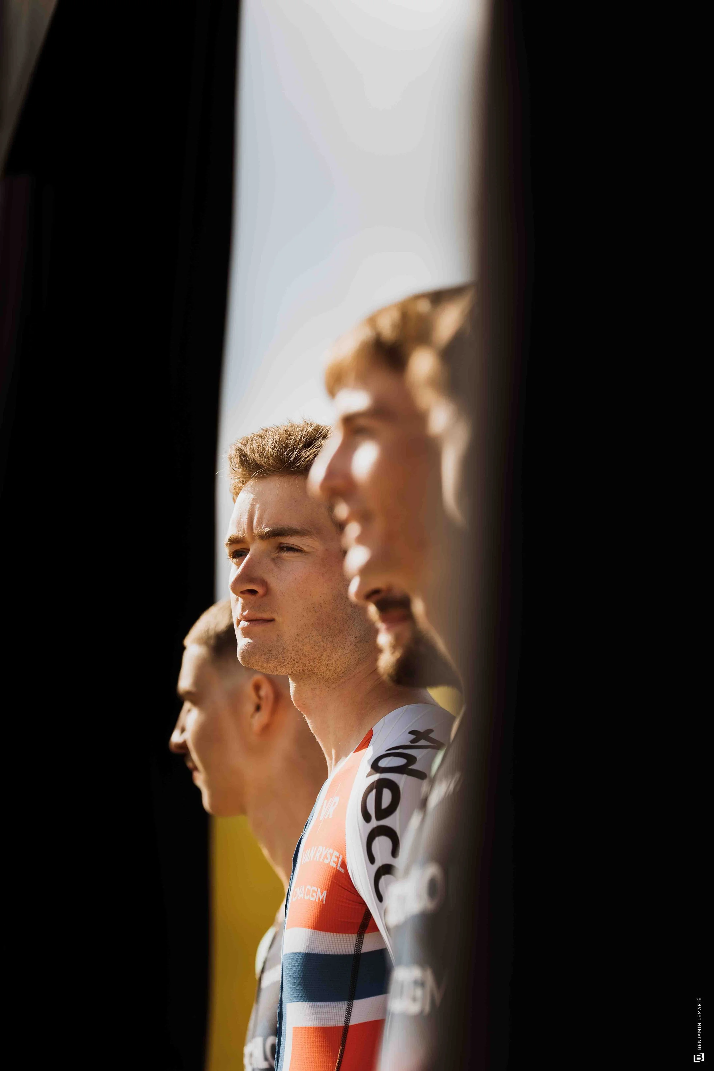 Une rangée de cyclistes professionnels alignés, portant des maillots de course, regardant droit devant, avec un focus sur le visage d'un homme au centre, en extérieur, sous la lumière naturelle.