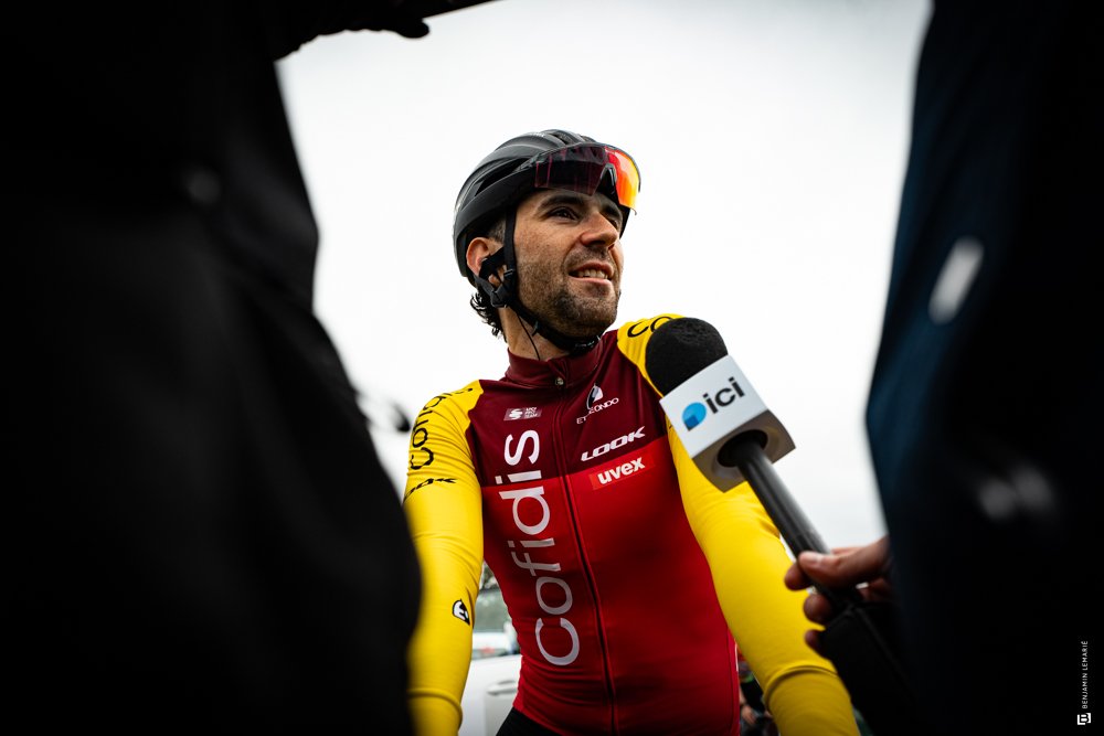 Un homme en tenue de cyclisme jaune et rouge parle à un microphone avec le logo d'« ici », face à la caméra, portant un casque de vélo, lors d'une interview.