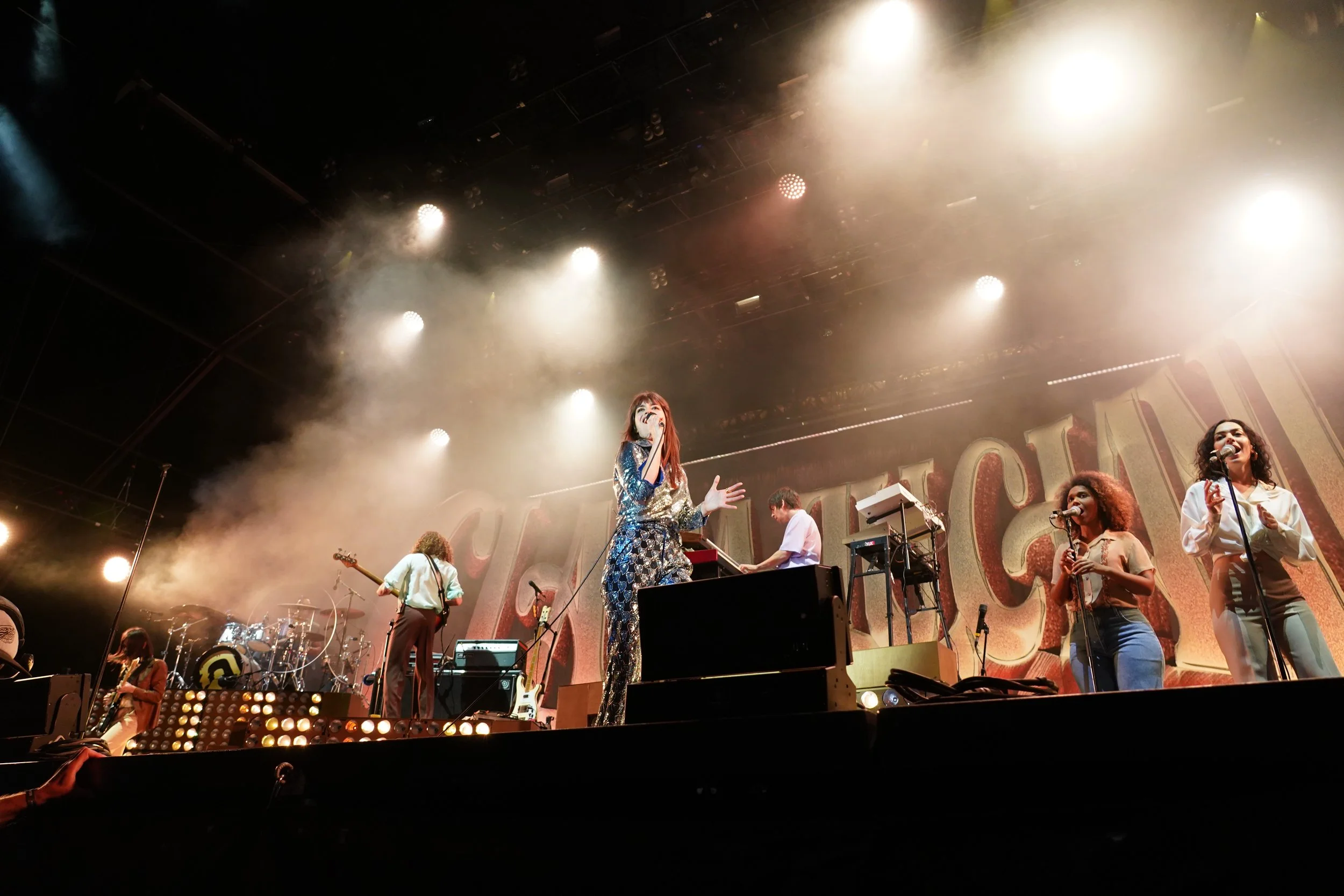 Une scène de concert avec des musiciens et chanteurs sur scène, éclairée par des projecteurs, ambiance musicale et lumière vive.