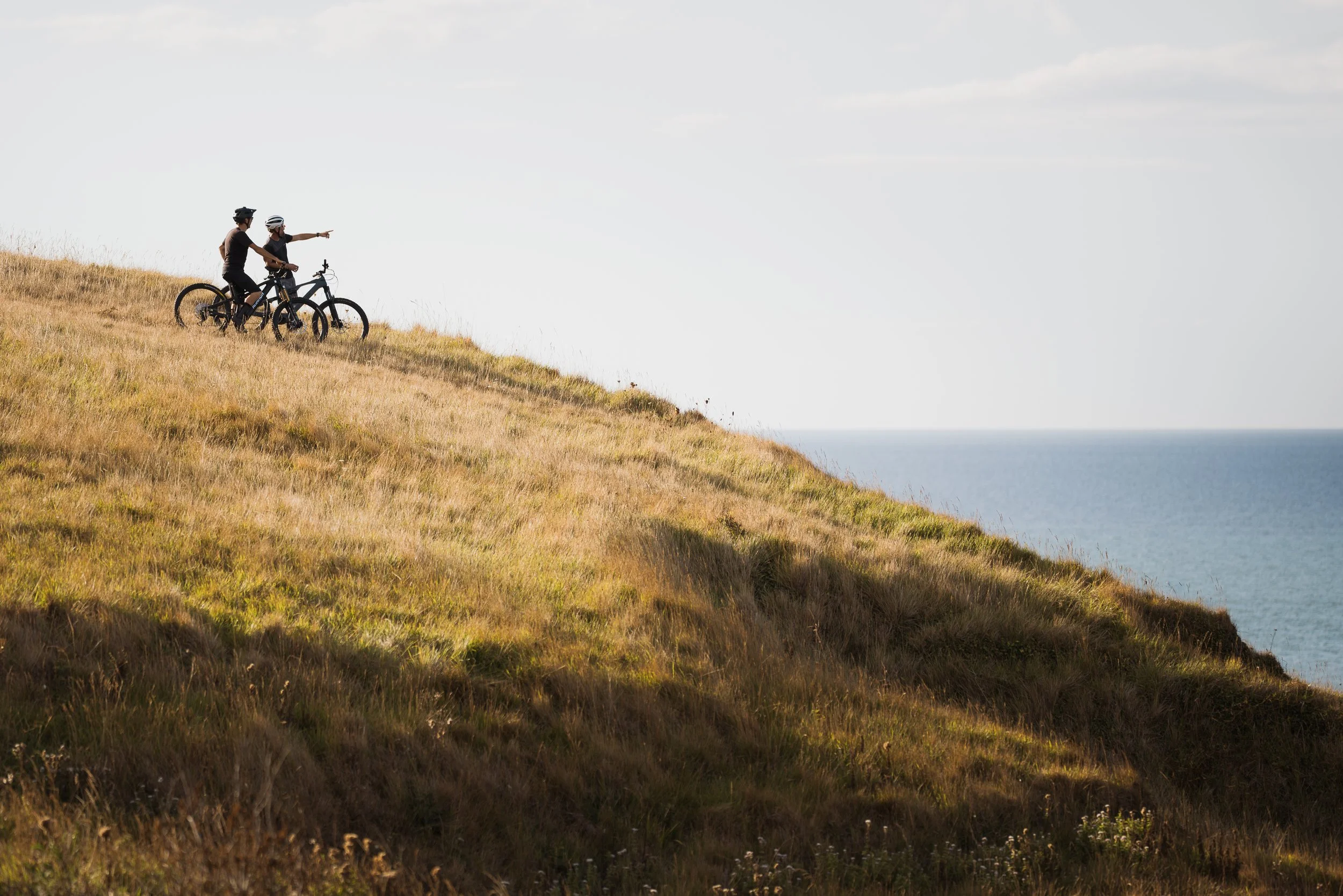 Deux personnes faisant du vélo en haut d'une colline, pointant vers l'horizon. Paysage vallonné avec l'océan en arrière-plan, ciel partiellement nuageux.