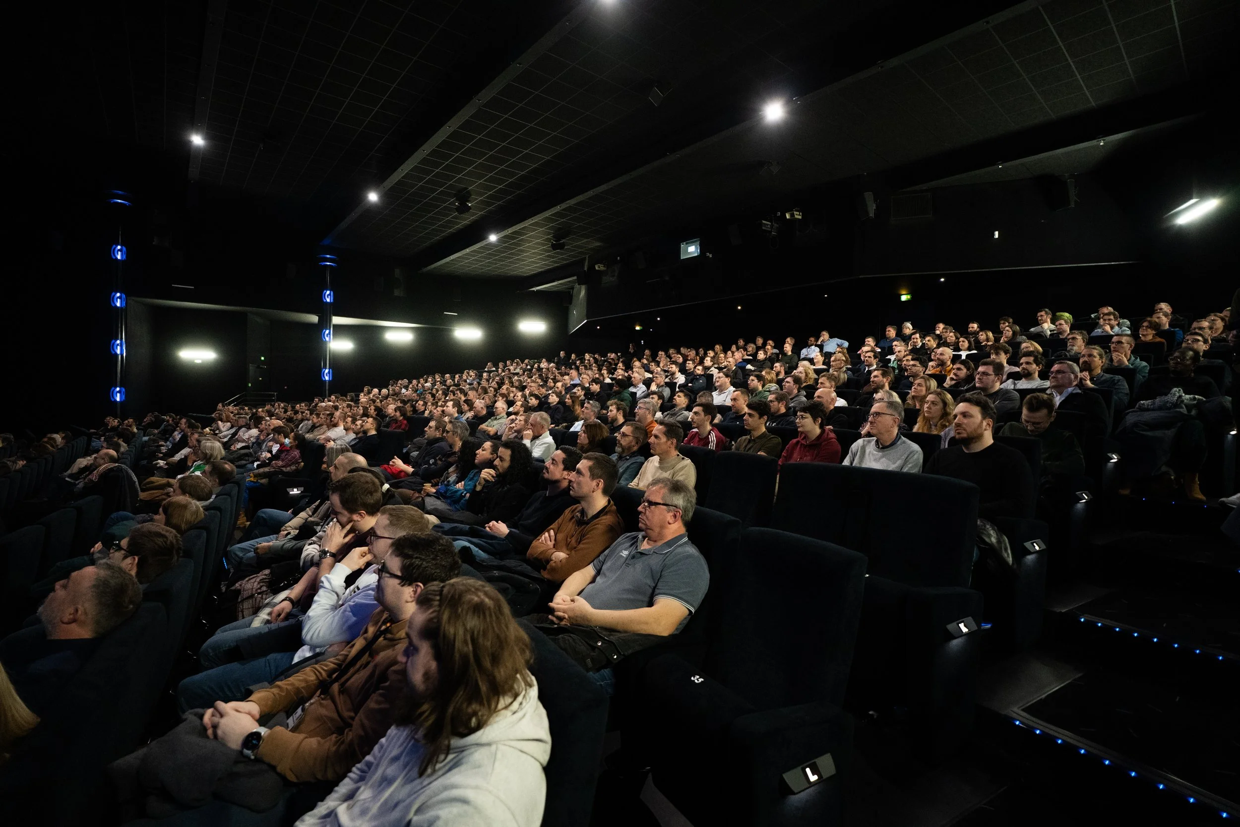 Une salle de conférence ou cinéma rempli de nombreuses personnes assises, regardant vers l'avant, dans un environnement sombre avec un plafond noir et des lumières discrètes.