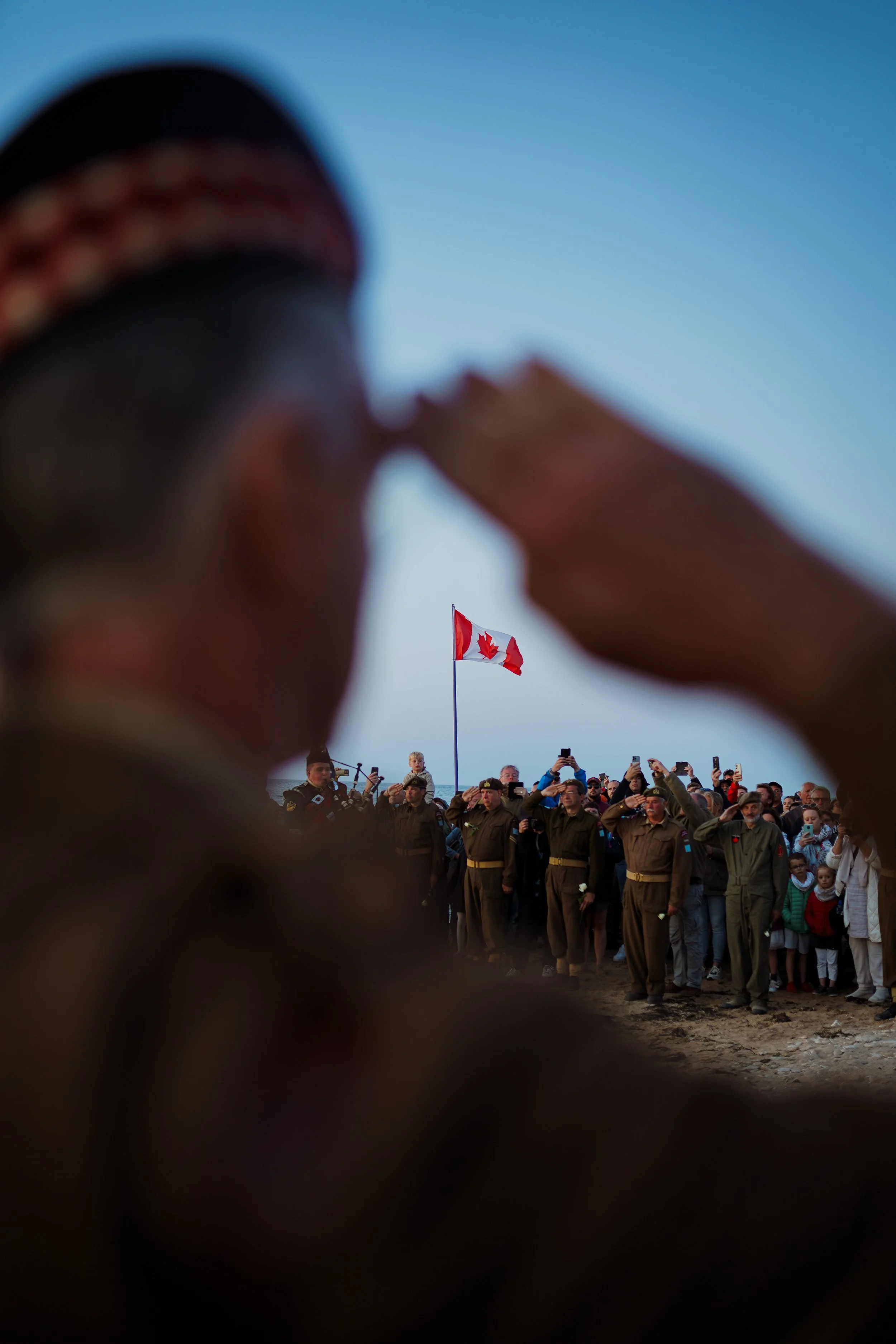 Un groupe de personnes, dont des policiers en uniforme, assistent à une cérémonie en plein air sous un ciel clair. À l'arrière-plan, un drapeau canadien est hissé. L'image est prise d'une perspective partiellement obstruée, probablement par une personne ou un objet proche de la caméra.