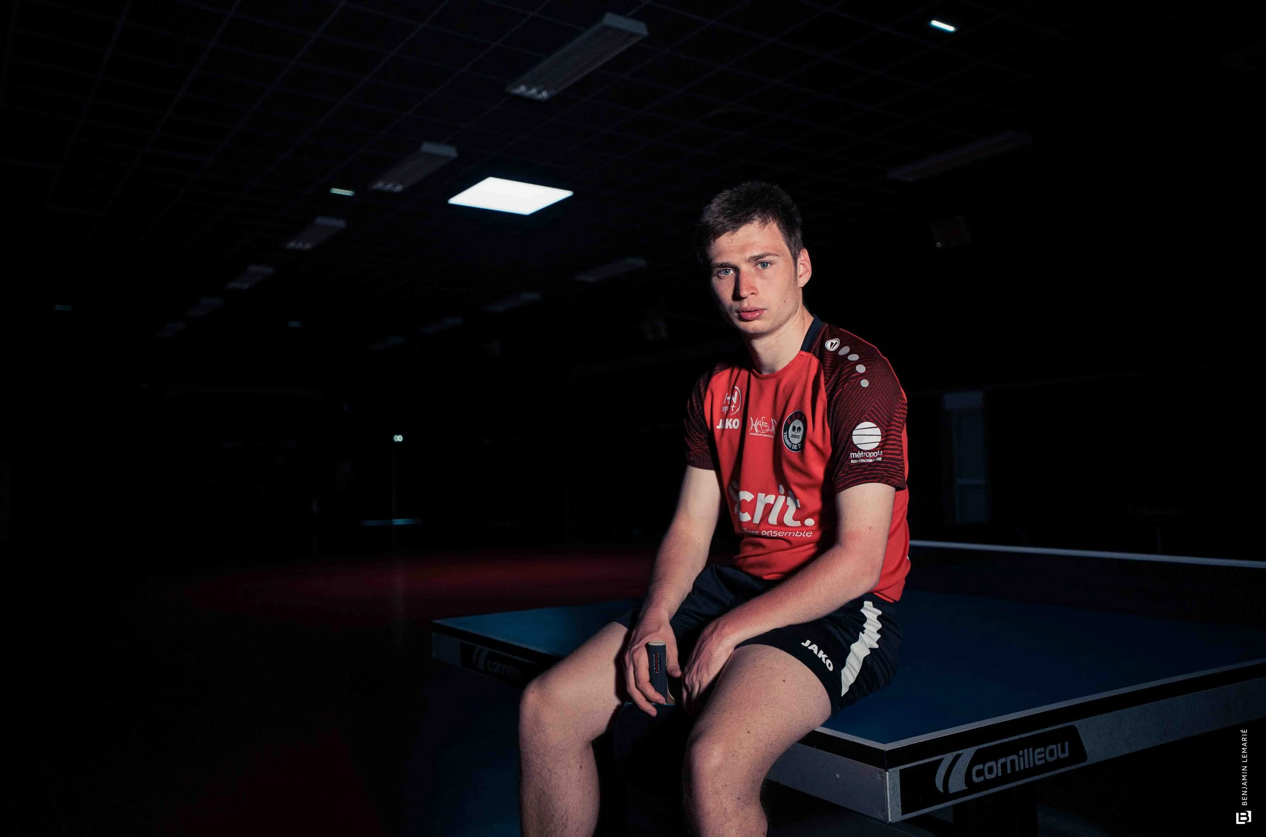 Un jeune homme portant un maillot de sport rouge assis sur une table de ping-pong dans une salle sombre, éclairé par un plafond lumineux.