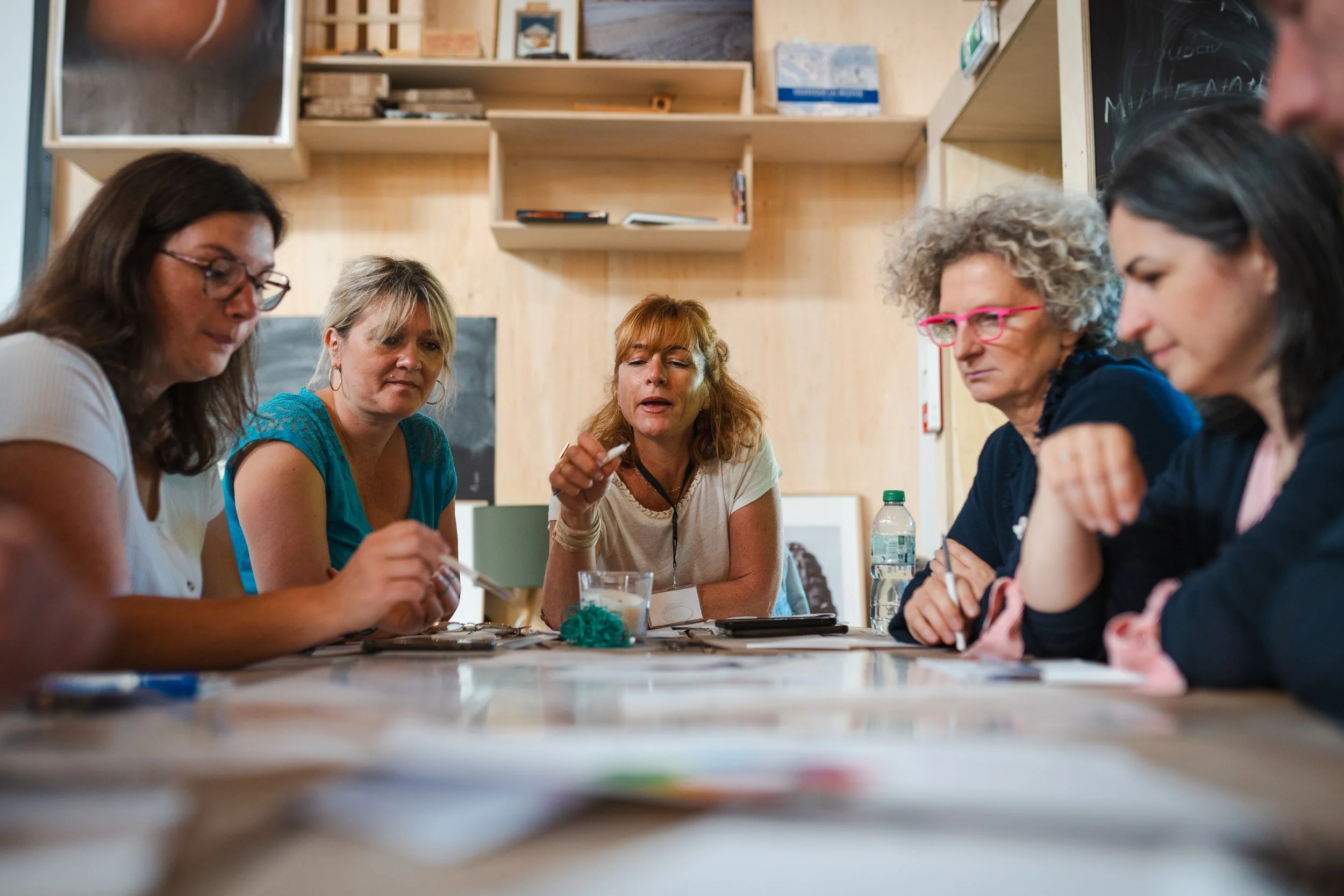 Groupe de cinq femmes assises autour d'une table en discussion dans une salle en bois. Certaines portent des lunettes et une bouteille d'eau est visible sur la table.