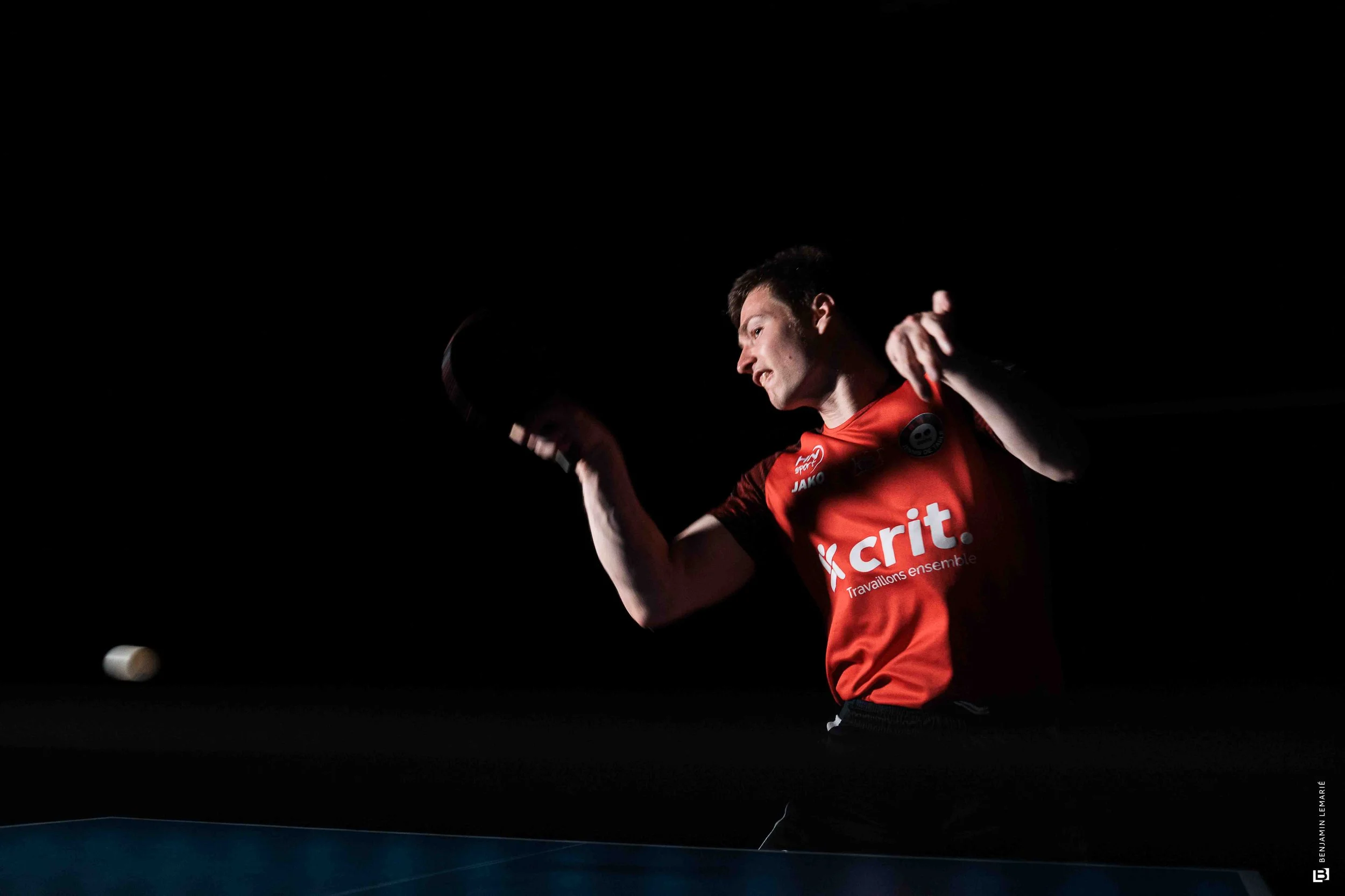 Un homme en tenue de sport rouge avec un logo 'crédit' joue au tennis de table dans une pièce sombre, seul éclairage sur lui.