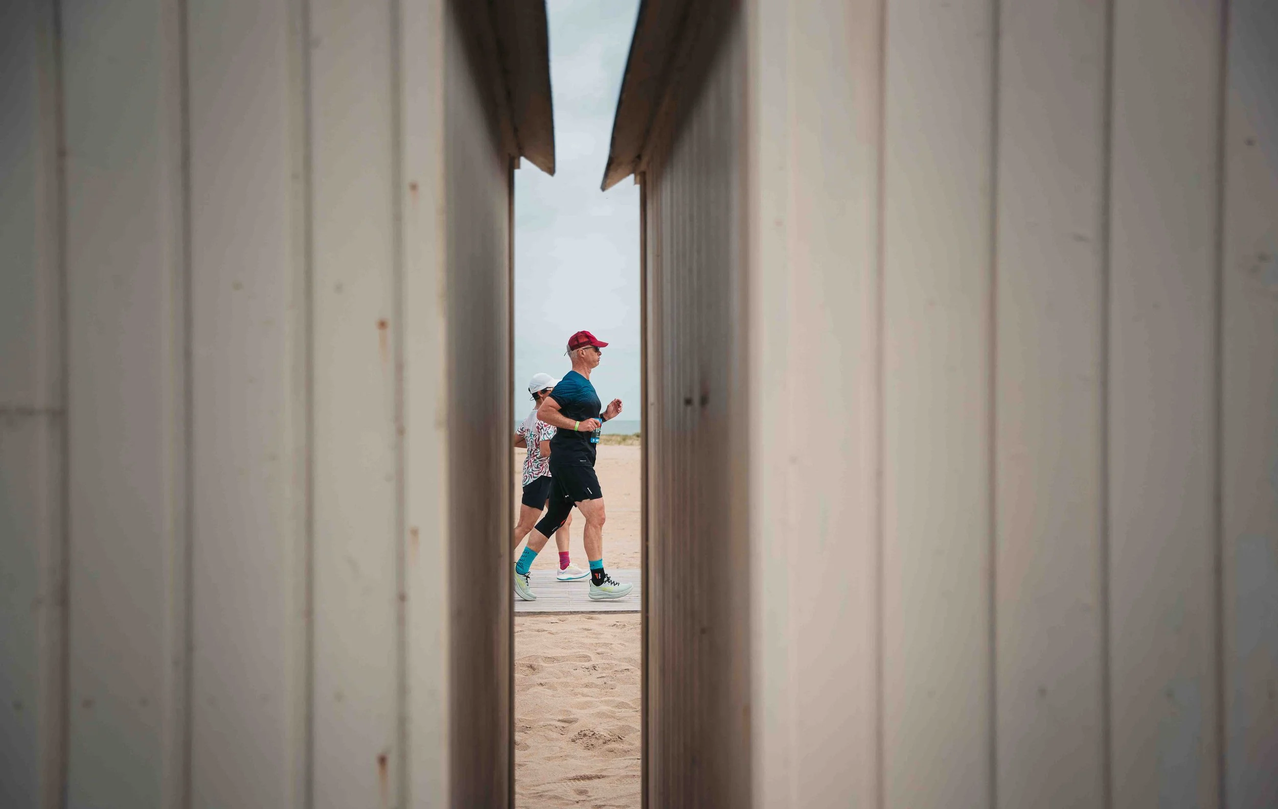 Deux personnes portant des vêtements de sport courent sur la plage vues à travers une ouverture entre deux planches en bois.