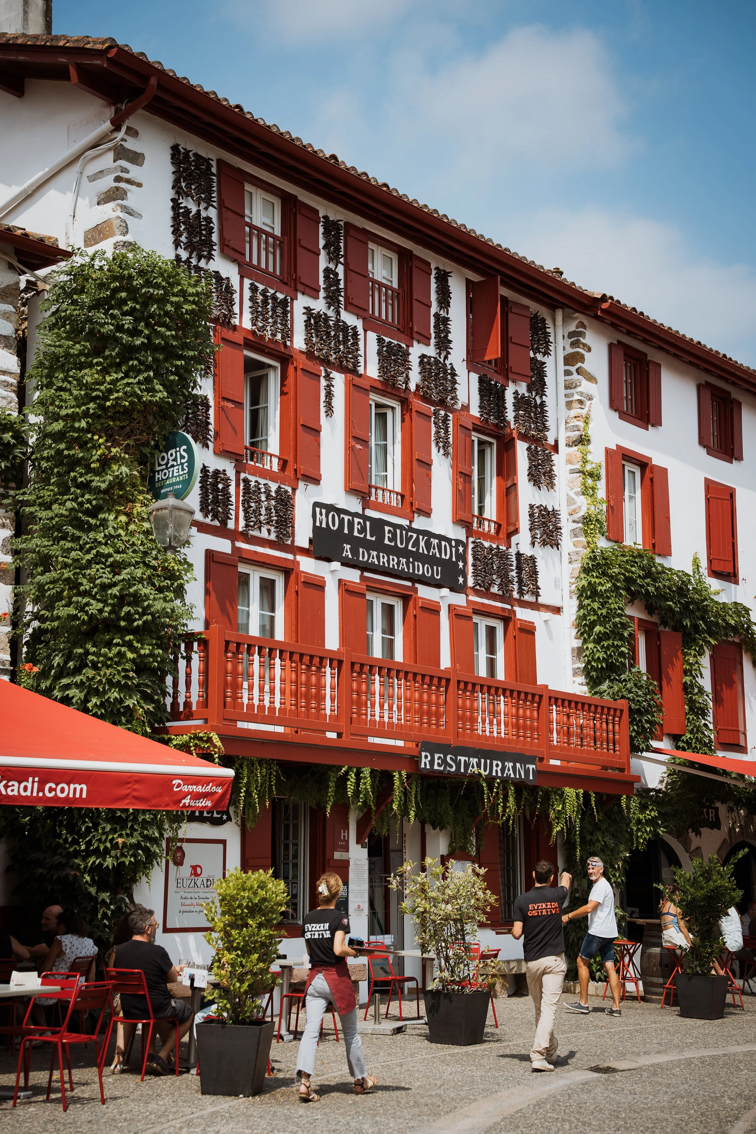 Façade d'un hôtel restaurant avec des volets rouges, décorations de piments séchés accrochés aux murs, terrasse extérieure avec des clients et des serveurs, panneaux indiquant le nom 'Motel Euzkadi' et 'Restaurant', un arbre vert et de petites plantes dans des pots