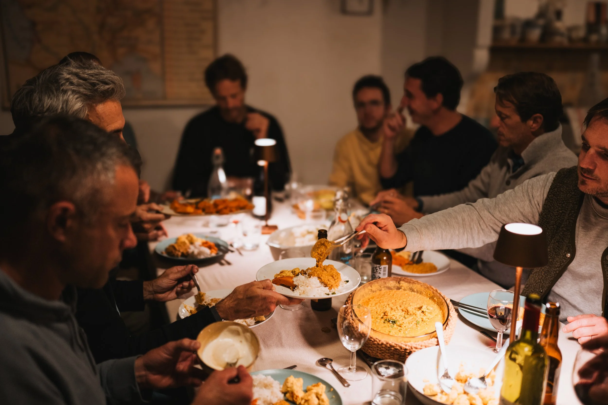 Groupe de personnes assises autour d'une table, partageant un repas à la maison dans une ambiance chaleureuse. Ils mangent du riz, du curry, et autres plats, avec des boissons, sous un éclairage tamisé.
