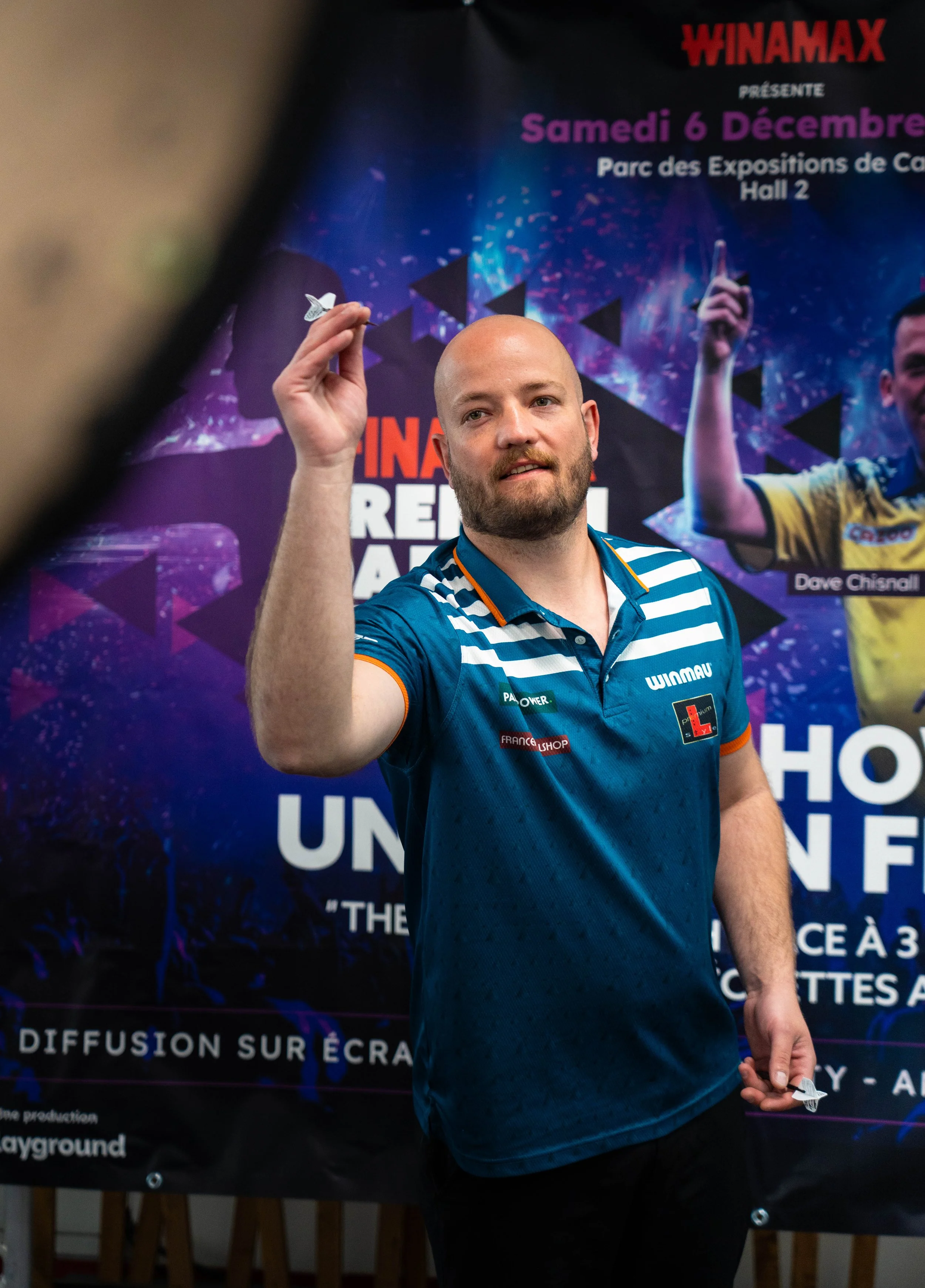 Homme en t-shirt bleu avec rayures blanches, tenant une fléchette dans sa main devant un affiche promotionnelle. L'affiche mentionne un événement le samedi 6 décembre au parc des expositions de Cannes.