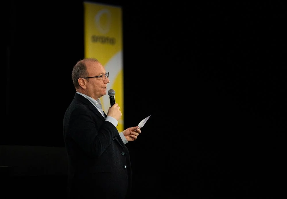 Un homme debout, portant des lunettes et un costume noir, tenant un micro et une feuille, en train de parler lors d'une présentation ou d'une conférence, avec un panneau jaune en arrière-plan.
