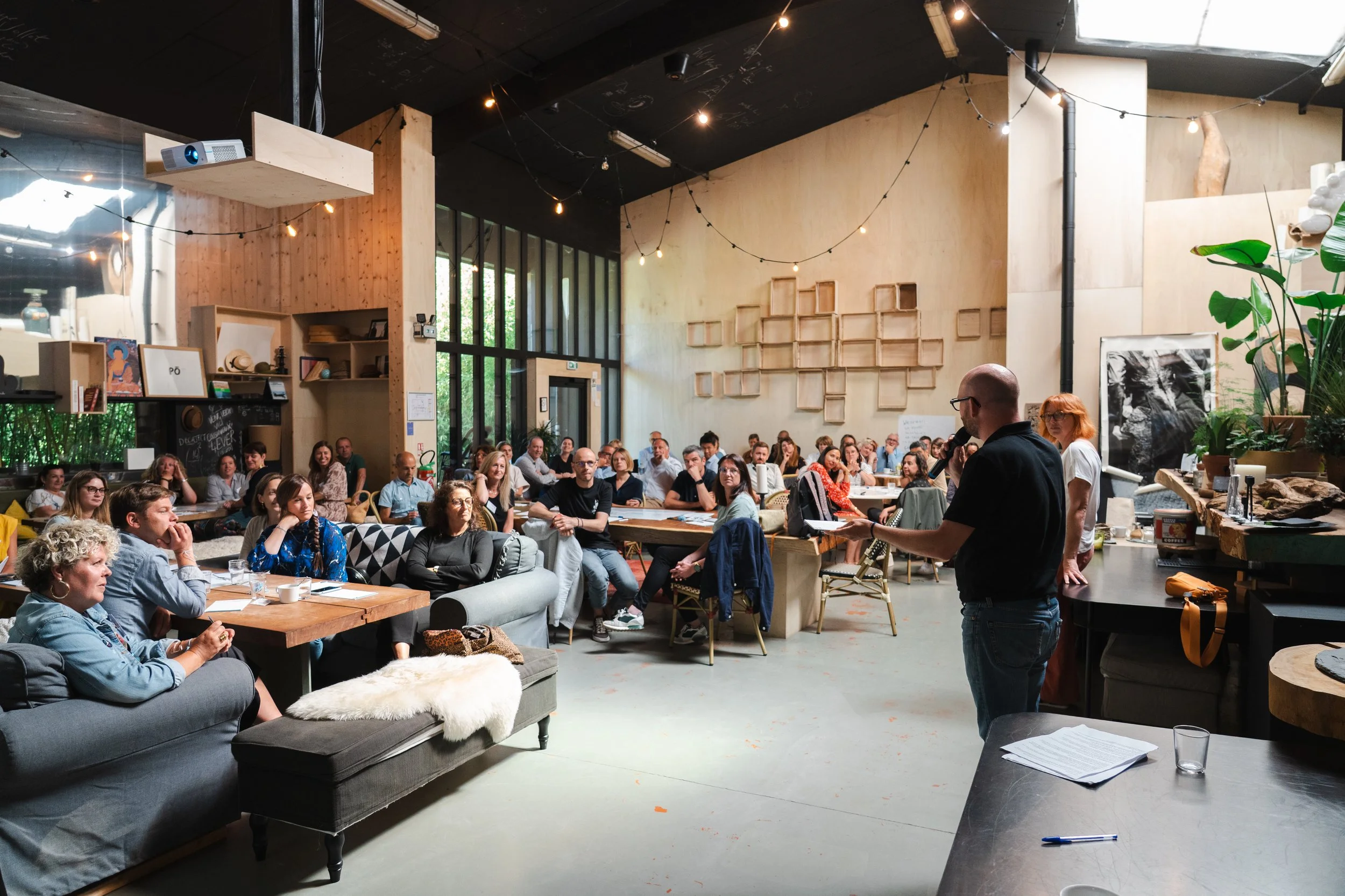 Une grande salle avec des personnes assises autour de tables, un homme avec un micro parle devant un groupe, décoration intérieure avec des plantes, des œuvres d'art et des étagères en bois.