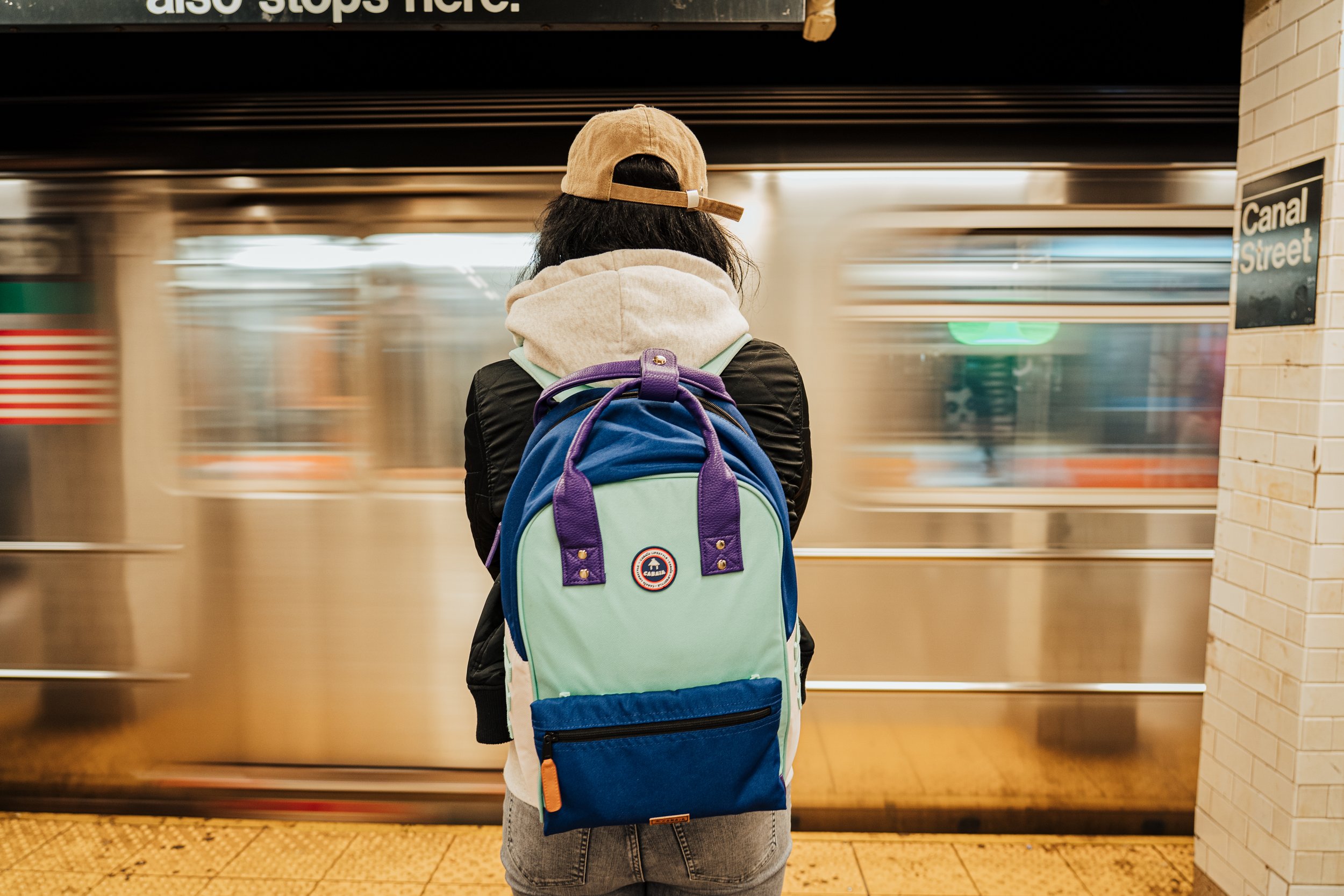 Une personne portant un sac à dos de couleur turquoise, vue de dos, attend devant un train en heure de pointe dans une station de métro. Elle porte une casquette beige et une veste noire, et se tient devant une porte de train métallique en mouvement.