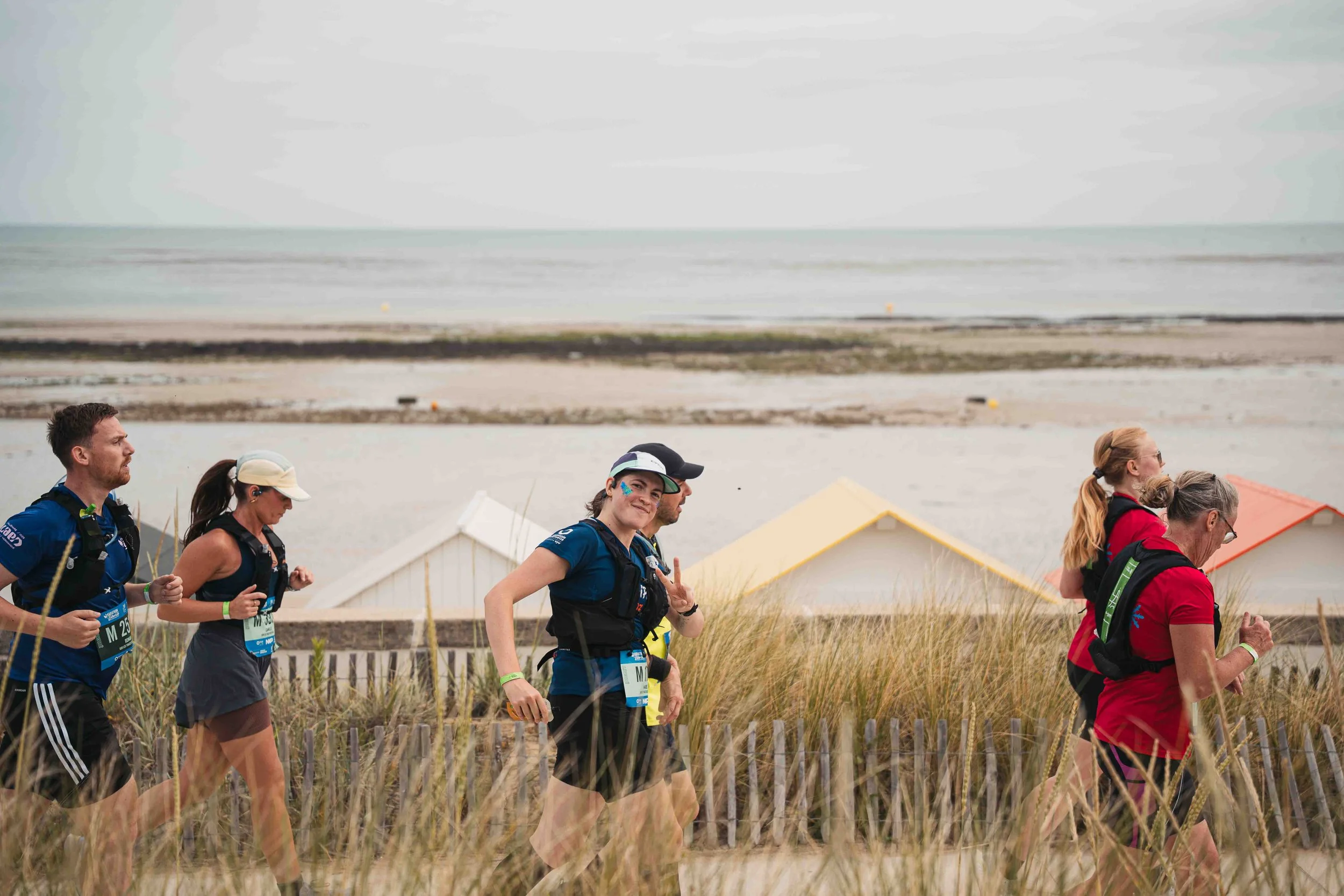 Groupe de personnes participant à une course ou une marche en extérieur près de la mer, avec des dunes et des maisons en arrière-plan.