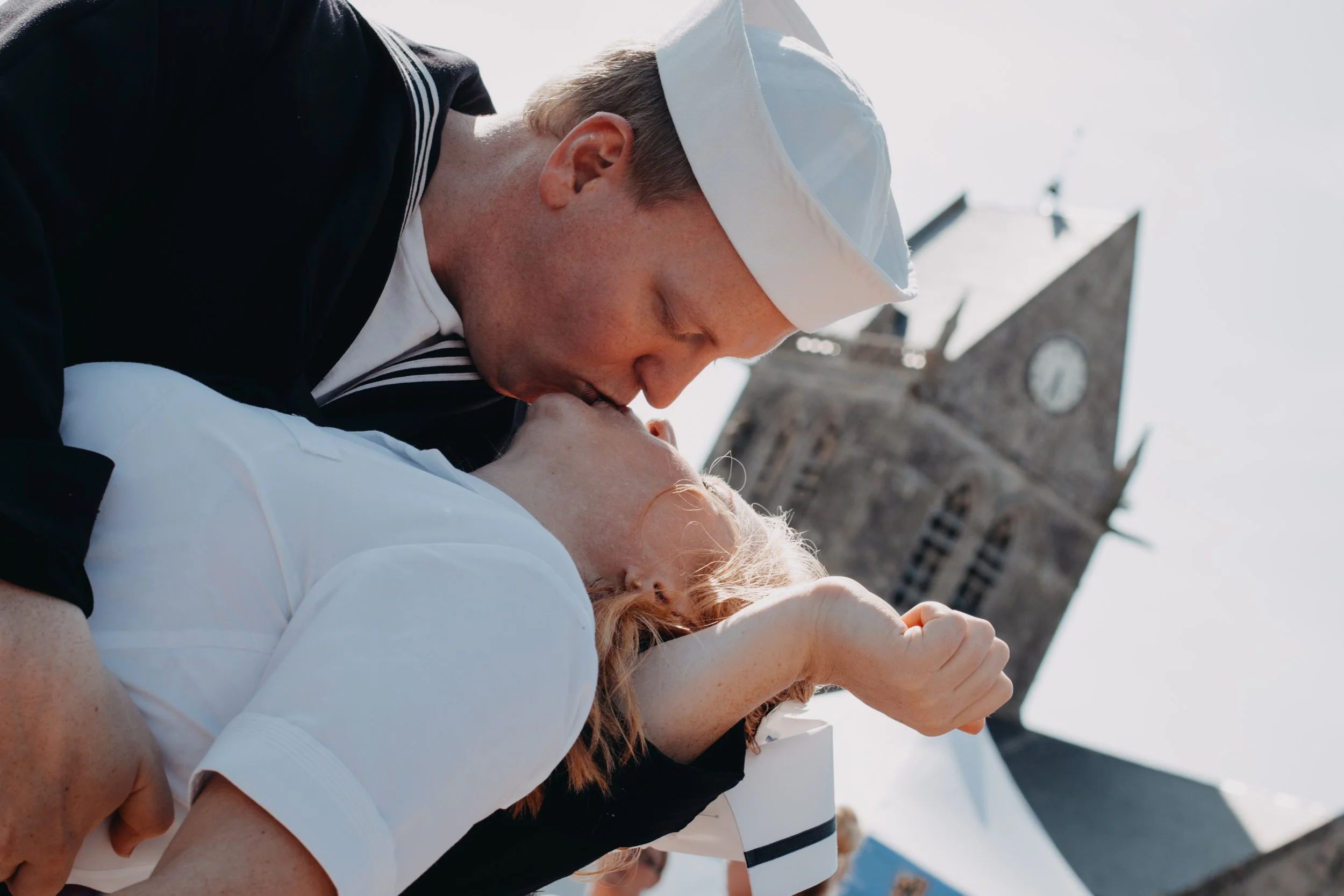 Un homme en uniforme marin embrasse une femme passionnément lors d'une cérémonie en plein air, avec une église en pierre en arrière-plan.