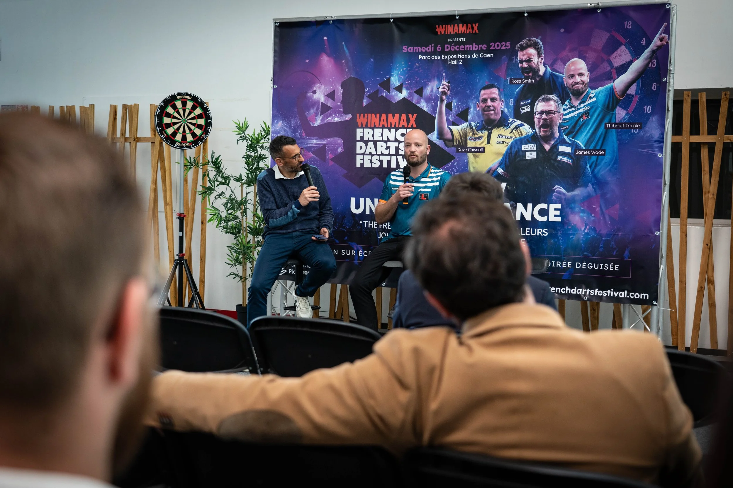 Une scène d'un festival de fléchettes avec deux hommes parlant dans des microphones devant une affiche promotionnelle du festival 'Winamax French Darts Festival', qui affiche la date, le lieu et des images de joueurs de fléchettes.