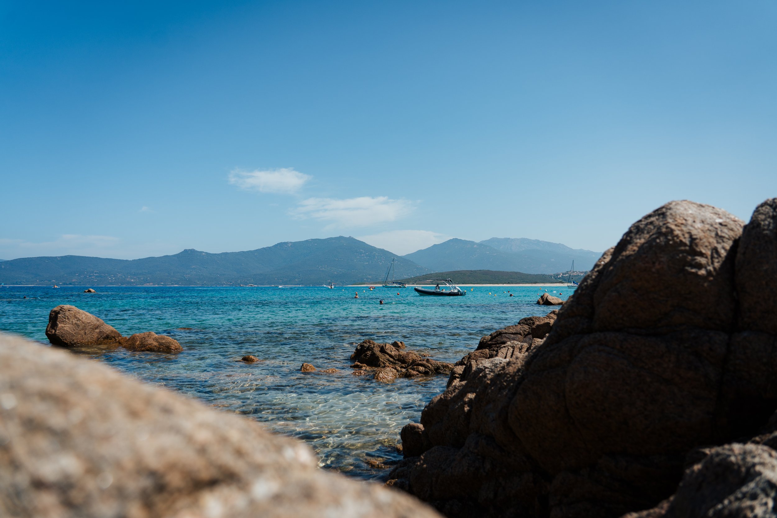 Plage avec rochers au premier plan, mer calme avec bateau à voile, montagnes en arrière-plan, ciel bleu avec quelques nuages.