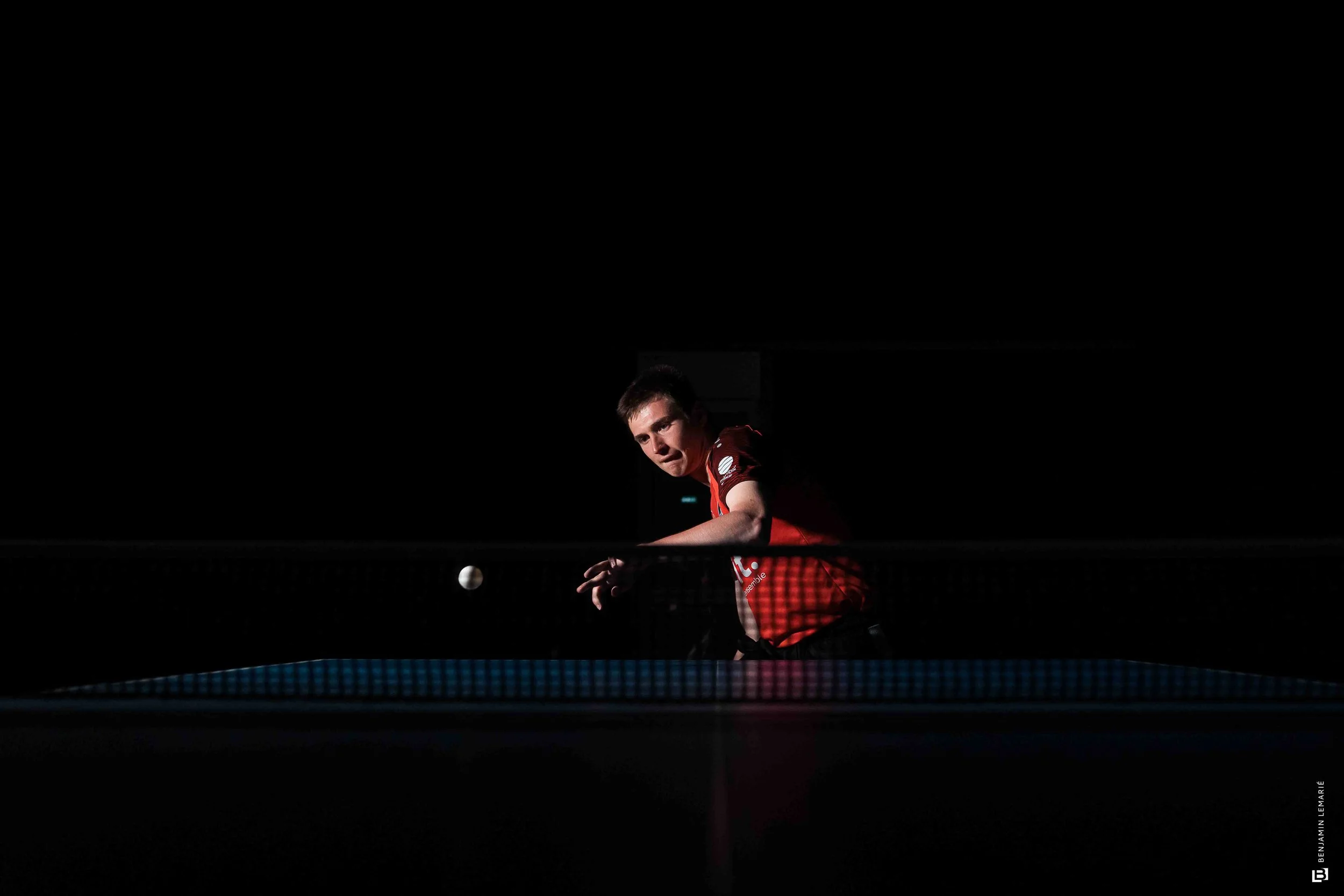 Un jeune homme joue au tennis de table dans une salle sombre, seul sous un éclairage ciblé. Il porte un maillot rouge. La balle blanche est en vol près de lui.