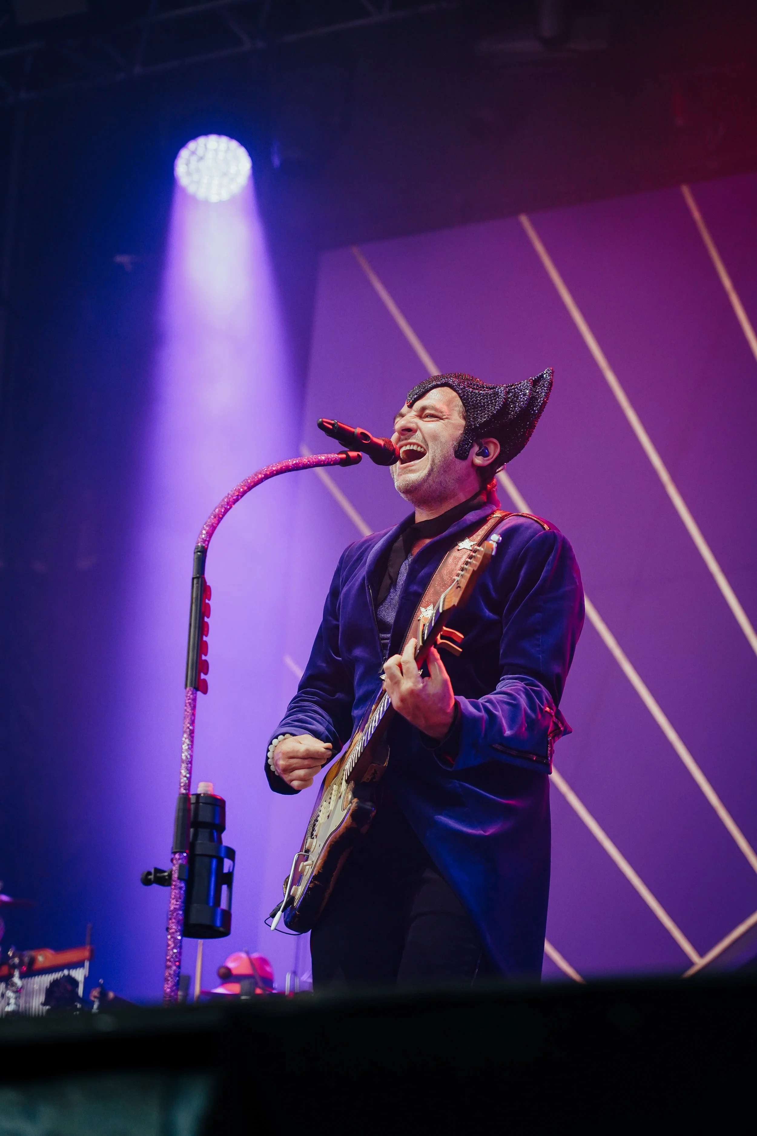 Un homme en costume noir chantant dans un micro, jouant de la guitare électrique sur scène avec des lumières violettes et un fond géométrique.