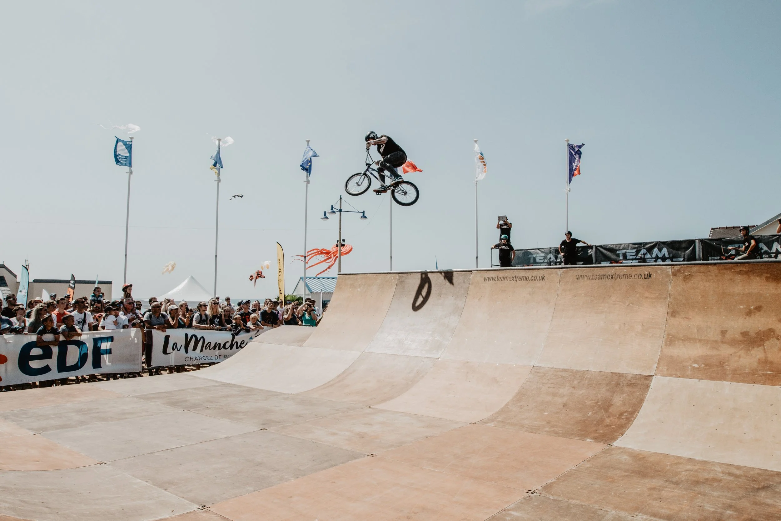Un athlète effectue une figure de BMX dans un skatepark, avec une foule de spectateurs en arrière-plan. Le ciel est clair et plusieurs drapeaux flottent.
