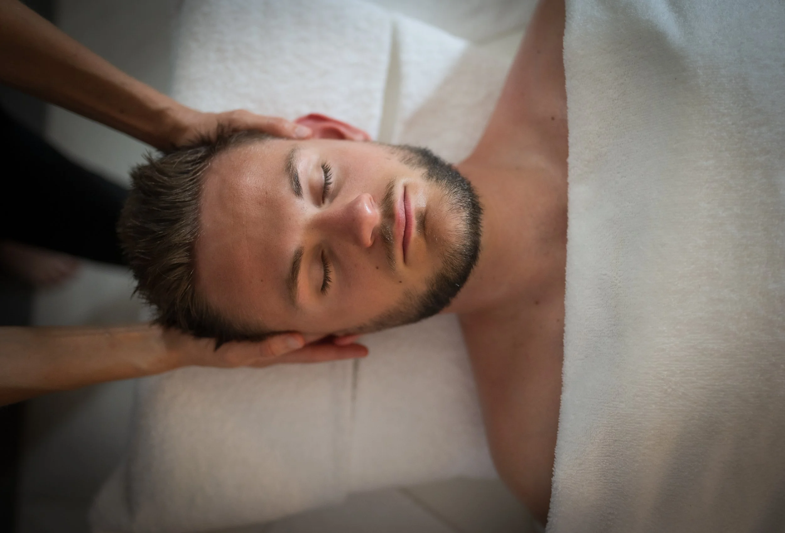 Homme recevant un massage à la tête, allongé sur une table avec une serviette blanche autour du corps.