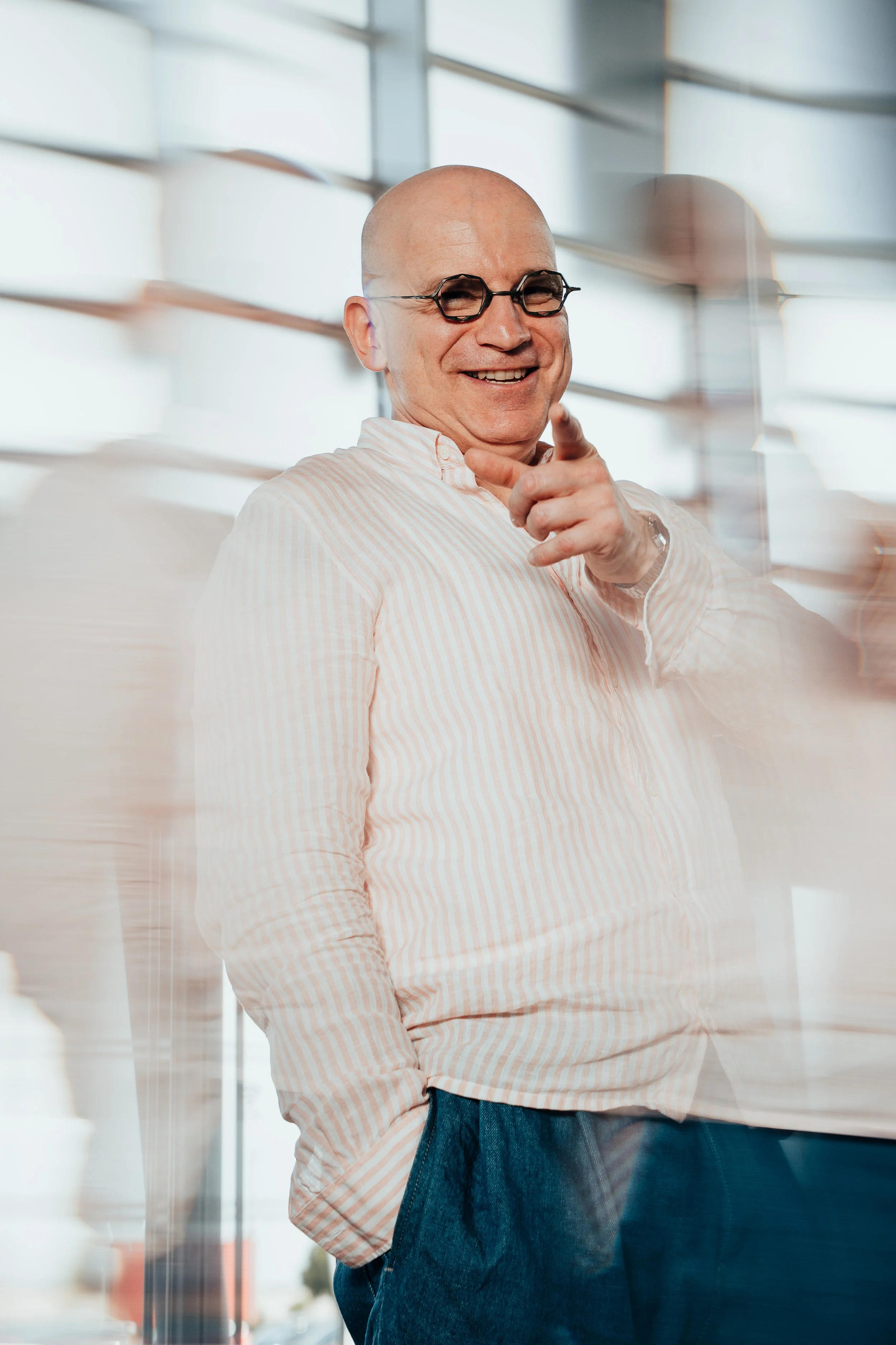 Un homme chauve portant des lunettes rondes et un chemisier à rayures roses et blanches, souriant et pointant vers l'avant dans un environnement lumineux.