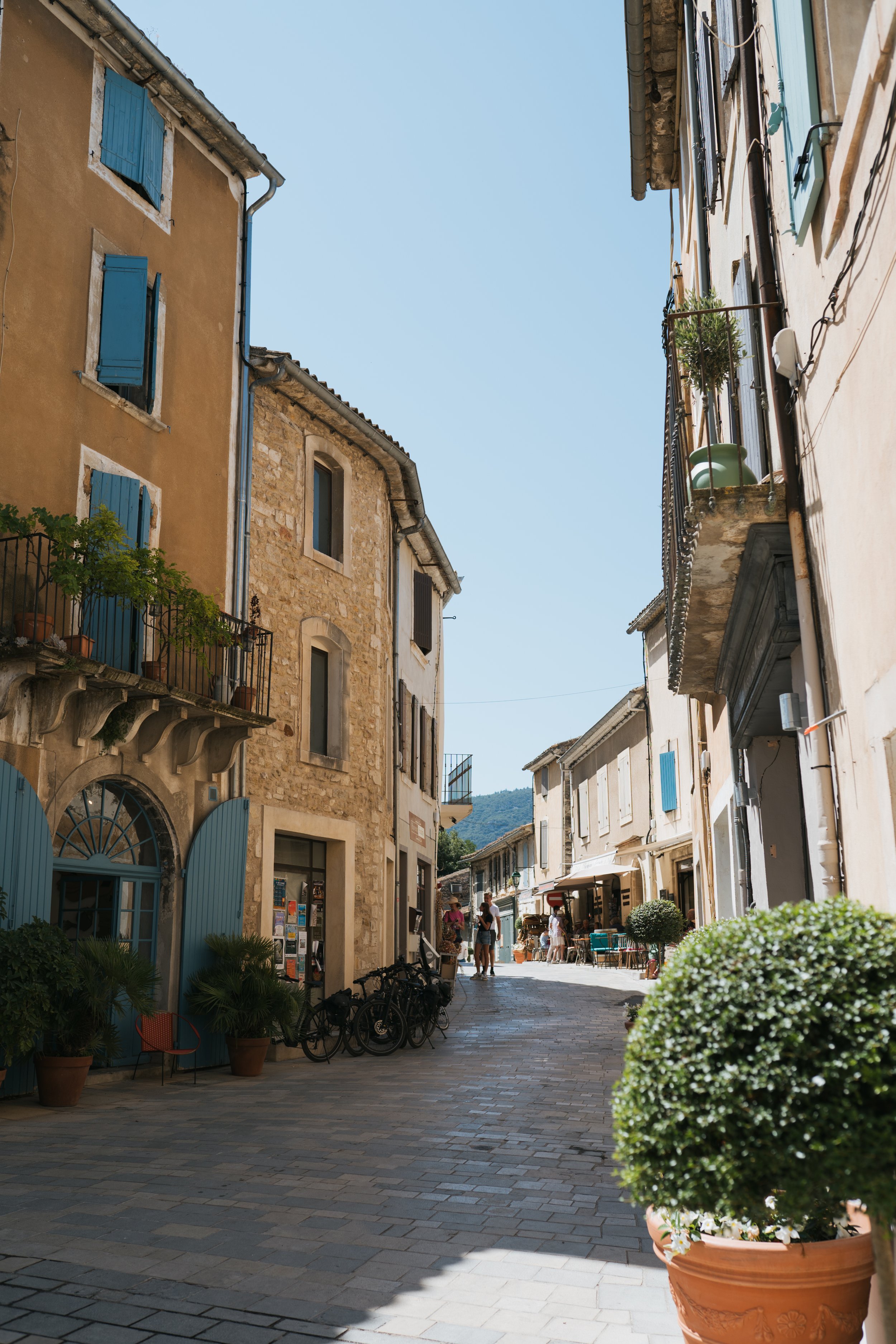 Rue piétonne dans une vieille ville, avec des bâtiments en pierre et en enduit coloré, terrasses de cafés, plantes en pots et quelques bicyclettes. Ciel clair avec peu de nuages, ambiance ensoleillée.