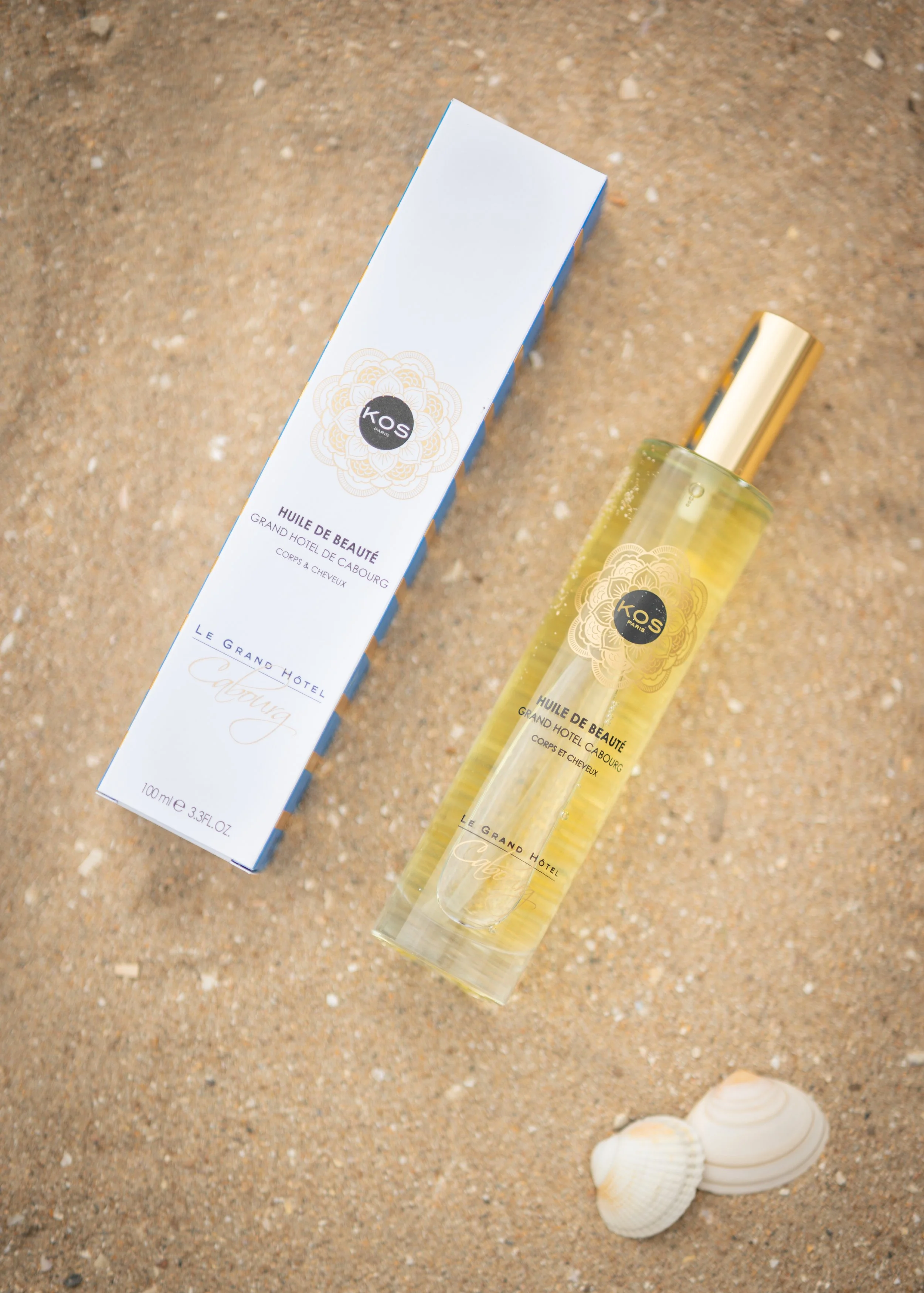 Un flacon de huile de beauté et sa boîte, ainsi que deux coquillages sur le sable.