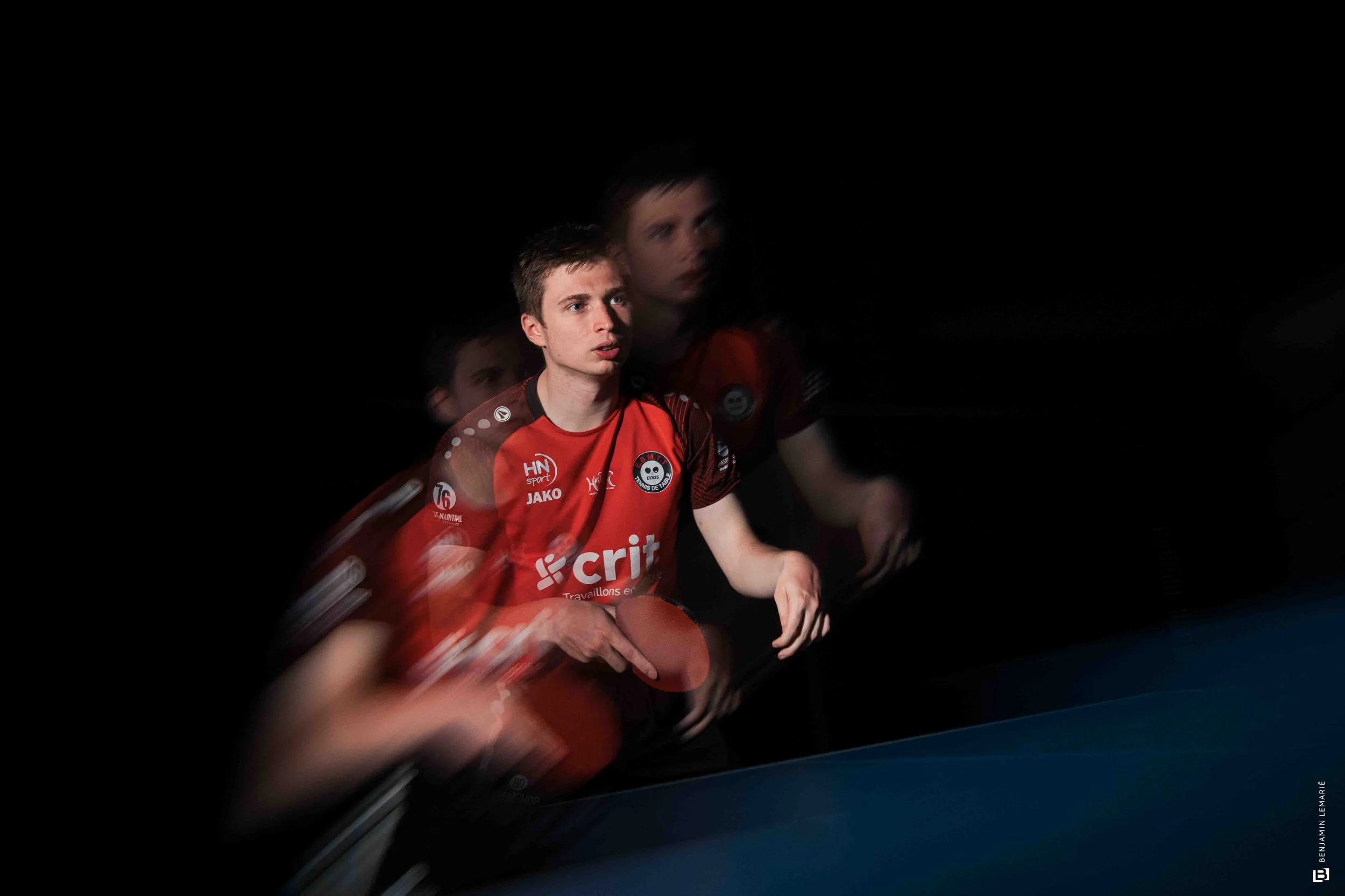 Deux jeunes hommes jouent au tennis en intérieur, l'un en premier plan avec un maillot rouge et le second en arrière-plan. La scène est sombre avec un éclairage concentré sur leur visage.