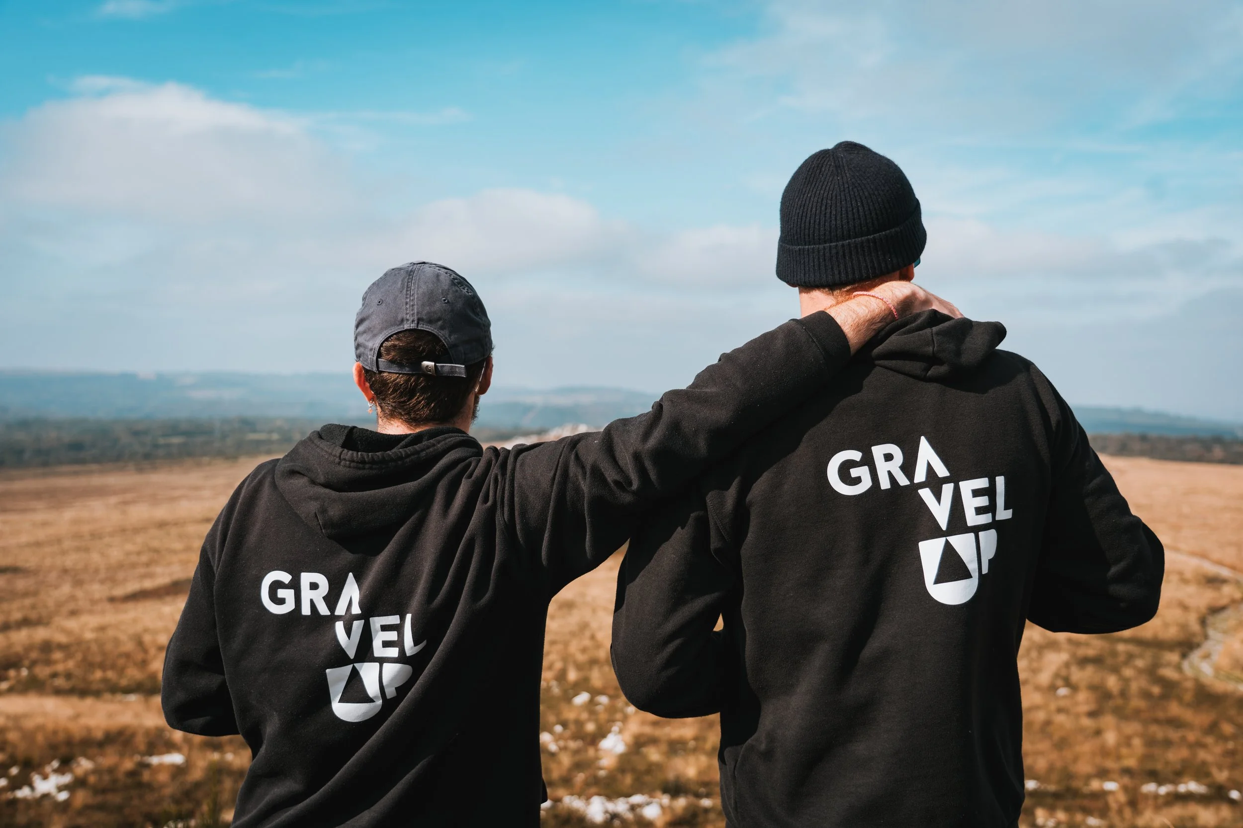 Deux personnes portant des sweatshirts noirs avec l'inscription "GRAVEL UP" à l'arrière, regardant au loin dans un paysage ouvert avec un ciel bleu clair.