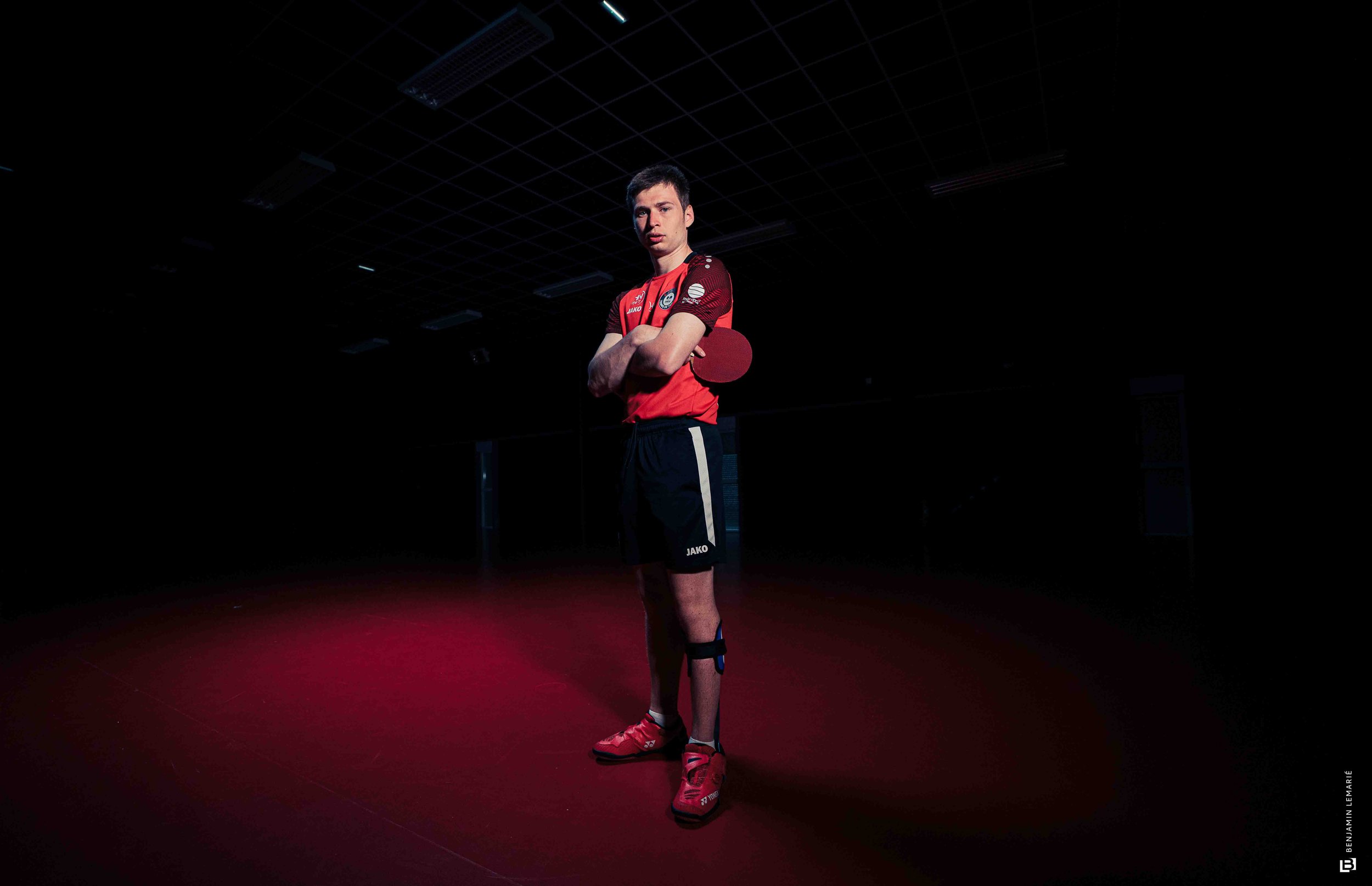 Jeune homme en tenue de ping-pong, debout dans une salle sombre, tenant une raquette de ping-pong, fond noir avec un plafond sombre.